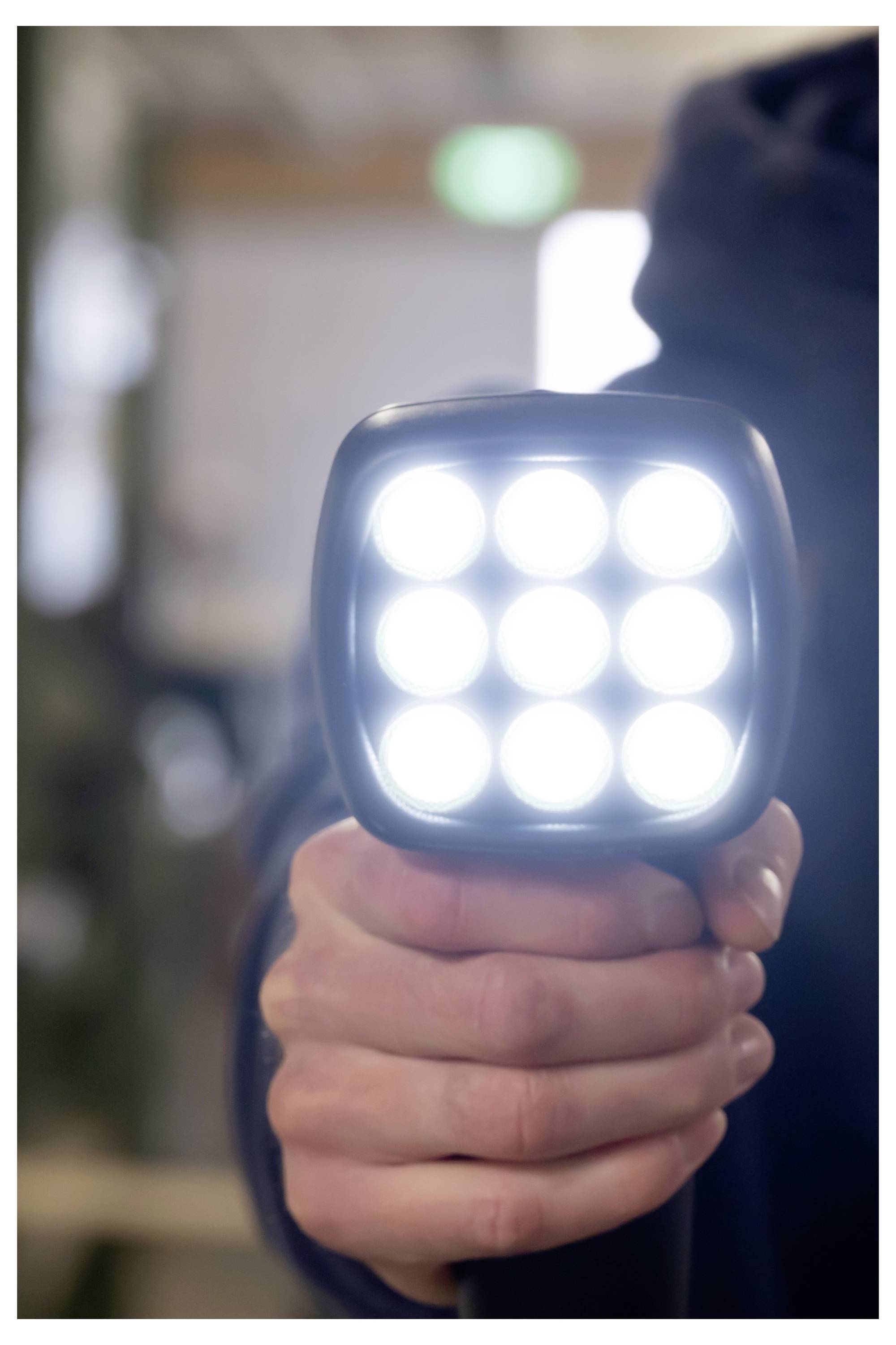 Nahaufnahme einer Hand, die eine helle quadratische LED-Taschenlampe oder Arbeitsleuchte hält und direkt in Richtung der Kamera leuchtet, in einem Innenraum.