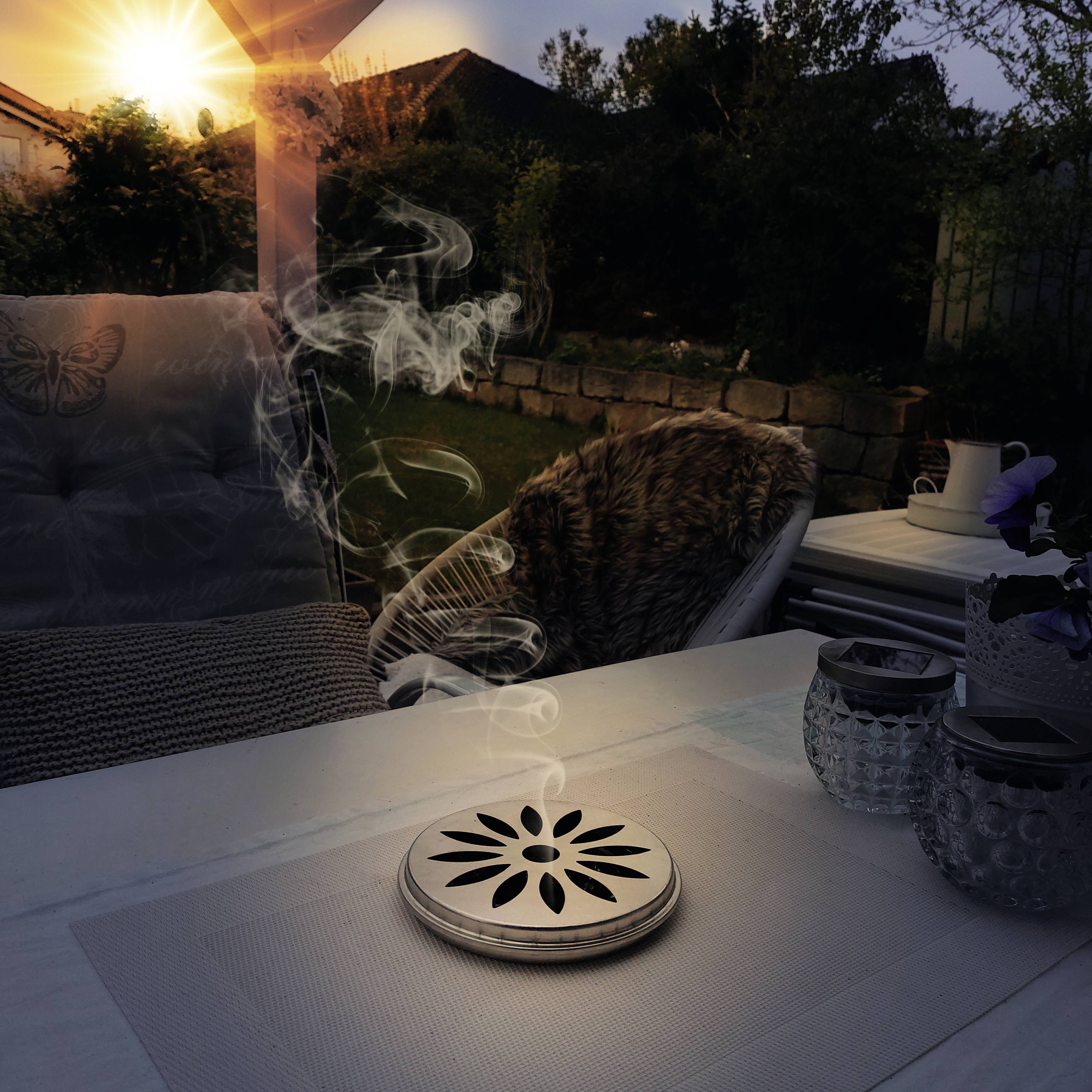 A cosy garden at sunset with a terrace, featuring a decorative smoke diffuser, surrounded by plants.