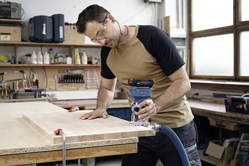 A man in safety glasses uses a handheld router on a wooden board in a workshop, showing woodworking activity and tool use.