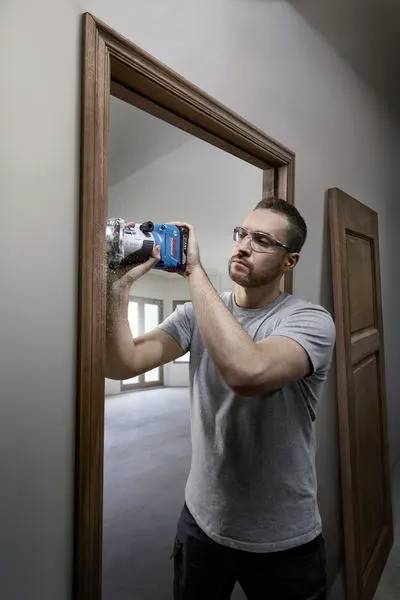 A man wearing protective glasses uses a power tool to work on a wooden doorframe inside a room.