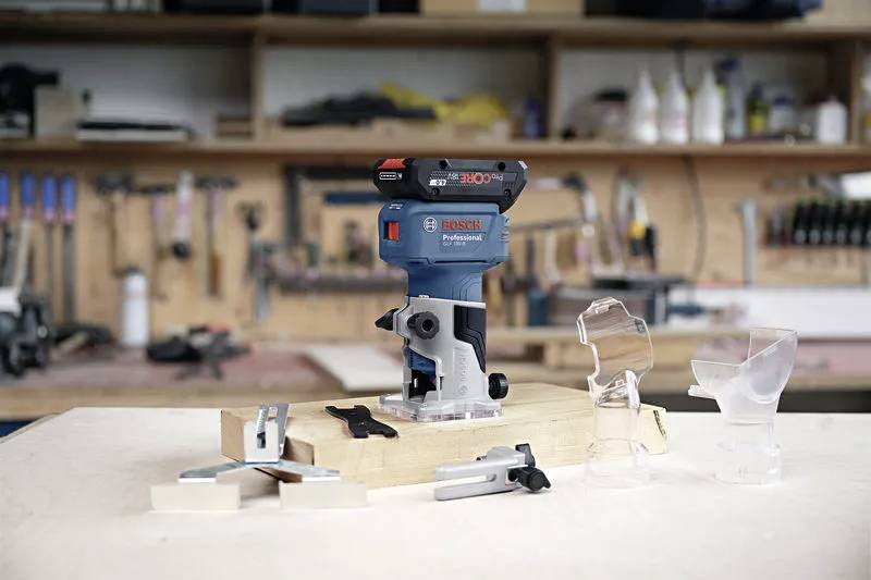 A compact blue and black woodworking router is placed on a workbench, surrounded by various transparent attachments and tools. A workshop setting is in the background.