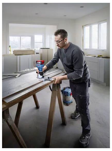 A man uses a power drill to assemble or repair a wooden worktable in a bright unfinished room.