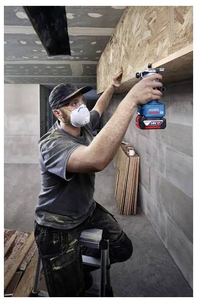 A masked worker uses a cordless drill to fasten a wooden panel overhead in an unfinished interior room.
