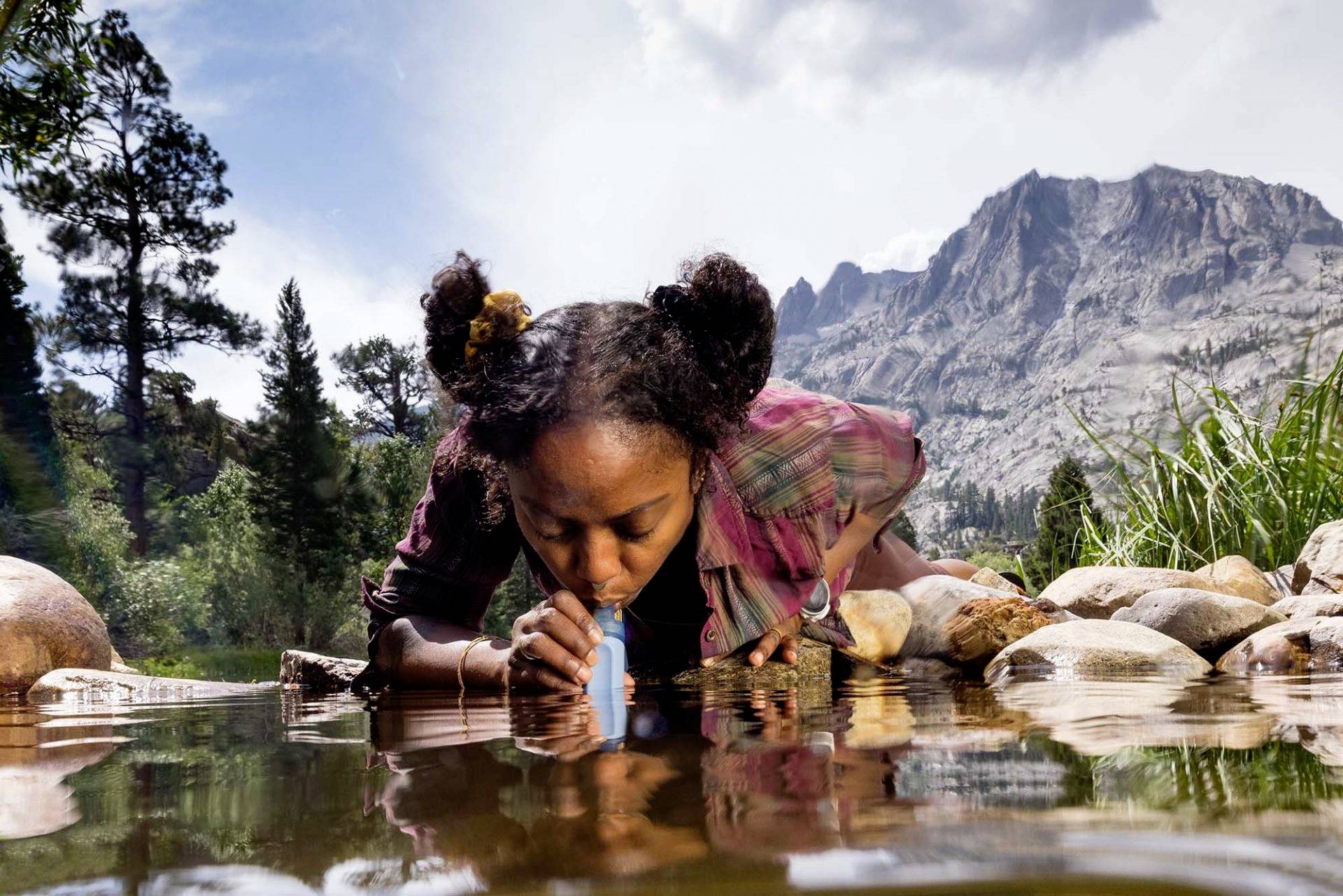 A person is drinking water from a stream in a mountainous landscape using a portable water filter. Mountains and trees are visible in the background.