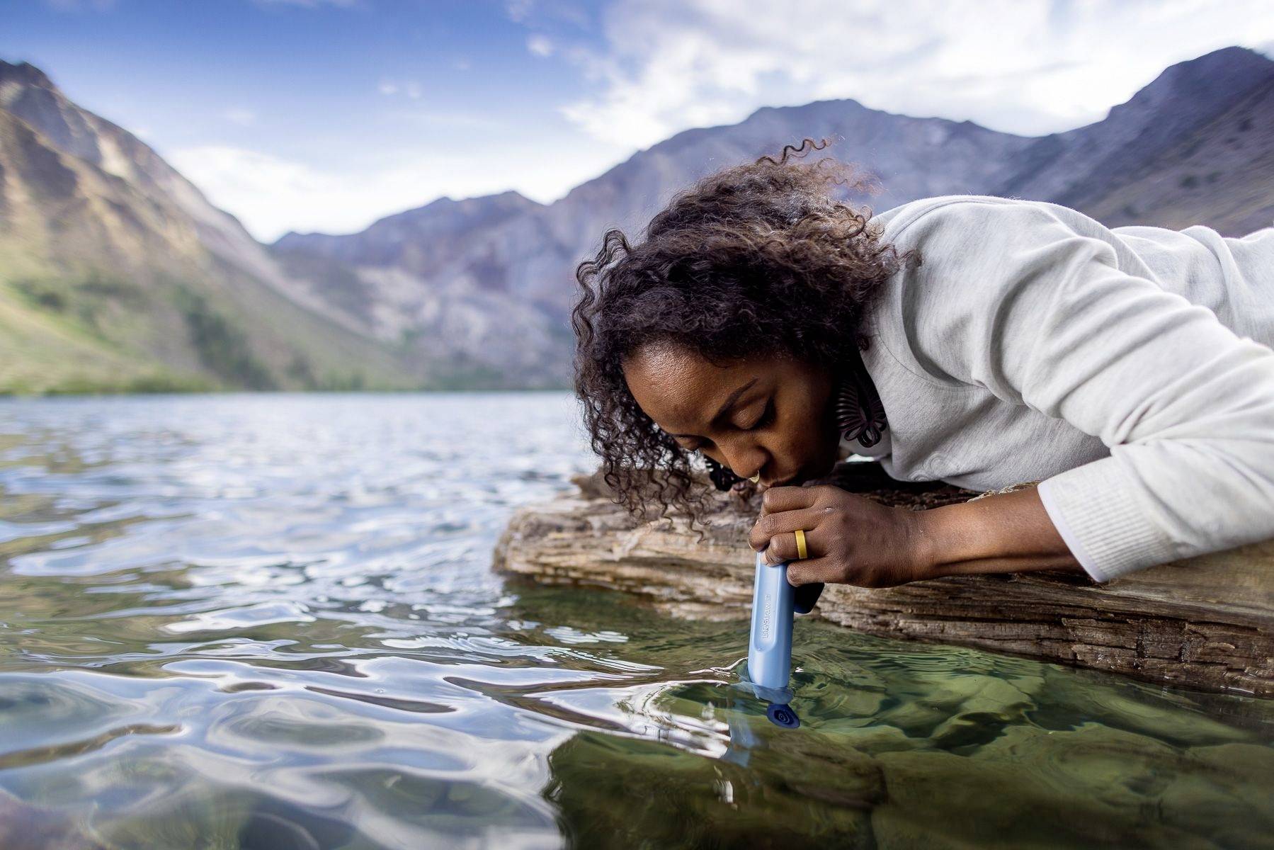 A person is drinking water from a clear mountain lake using a portable water filter. Mountains are visible in the background.