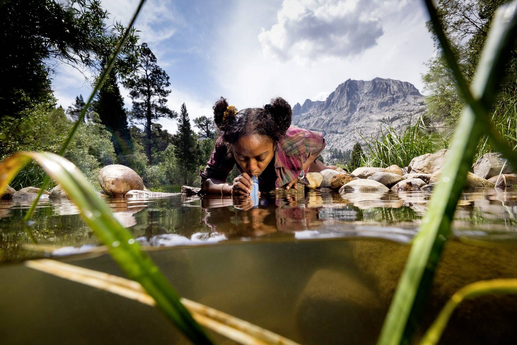 A person is drinking water from a stream in a mountainous landscape, surrounded by trees and rocks, beneath a cloudy sky.