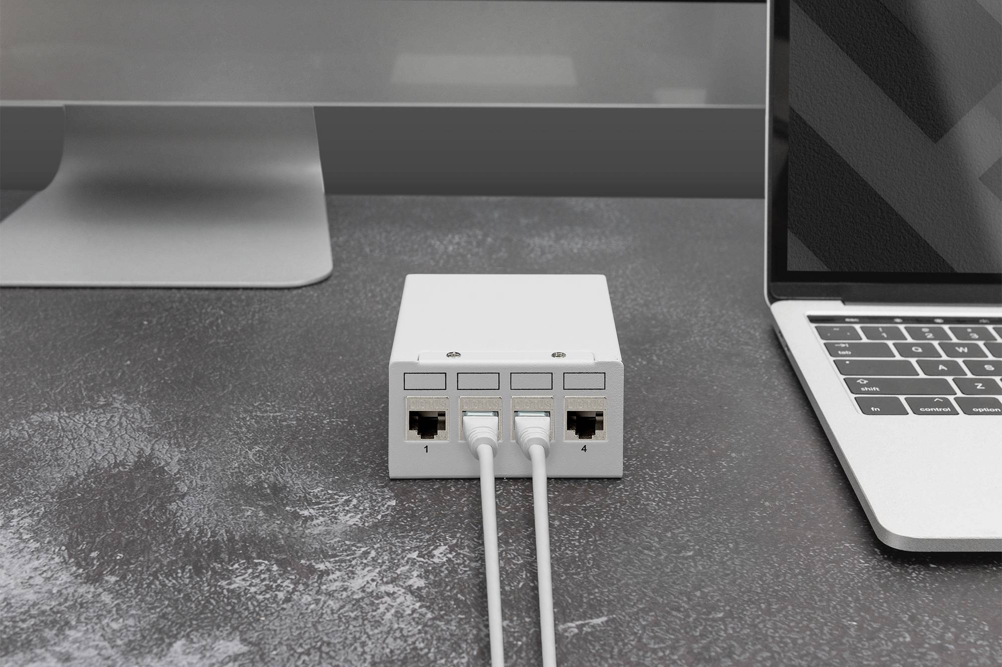 A small, white network switch connects two computers with network cables. The devices are situated on a grey desk.