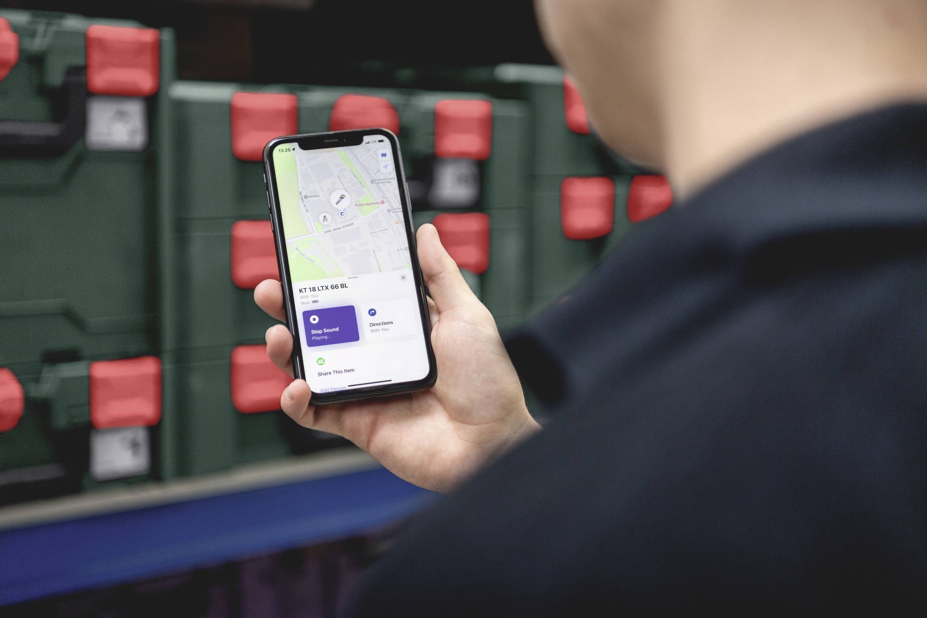 A person is holding a smartphone with an open navigation app. In the background, green boxes with red fastenings are visible.
