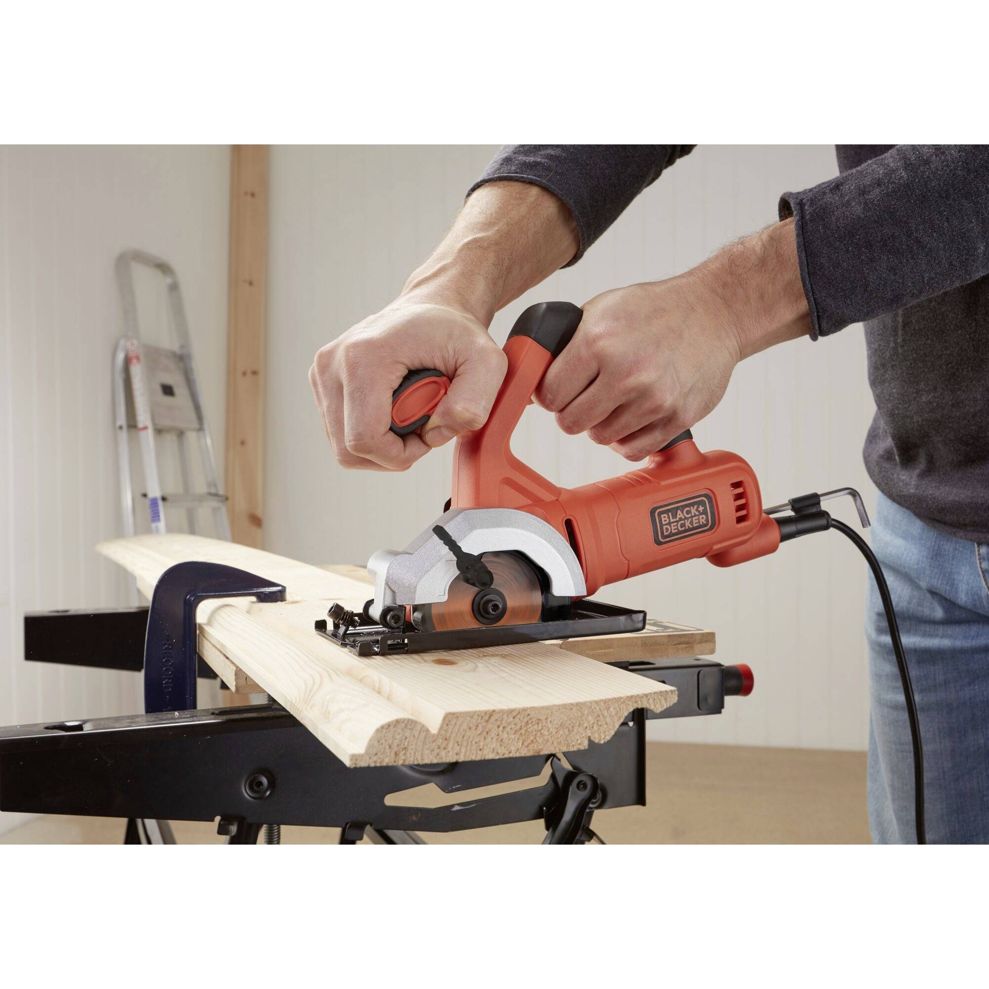 A person is sawing a wooden board with a red electric saw on a workbench. A ladder is standing in the background.