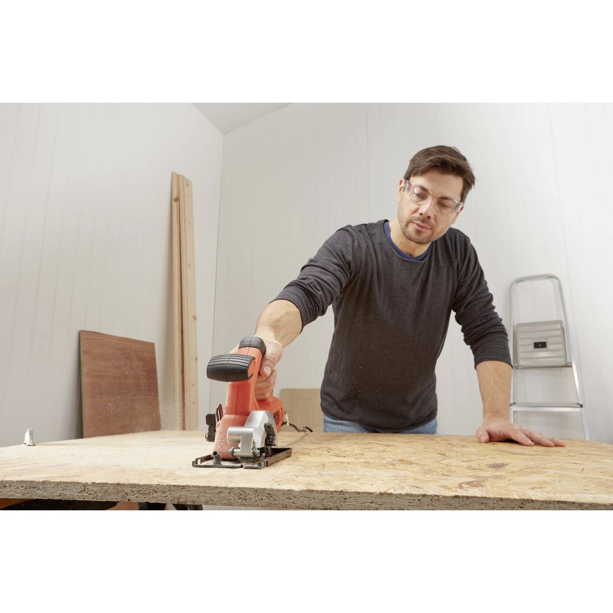 A middle-aged man is working with an electric saw on a wooden board in a workshop. A ladder is standing in the background.