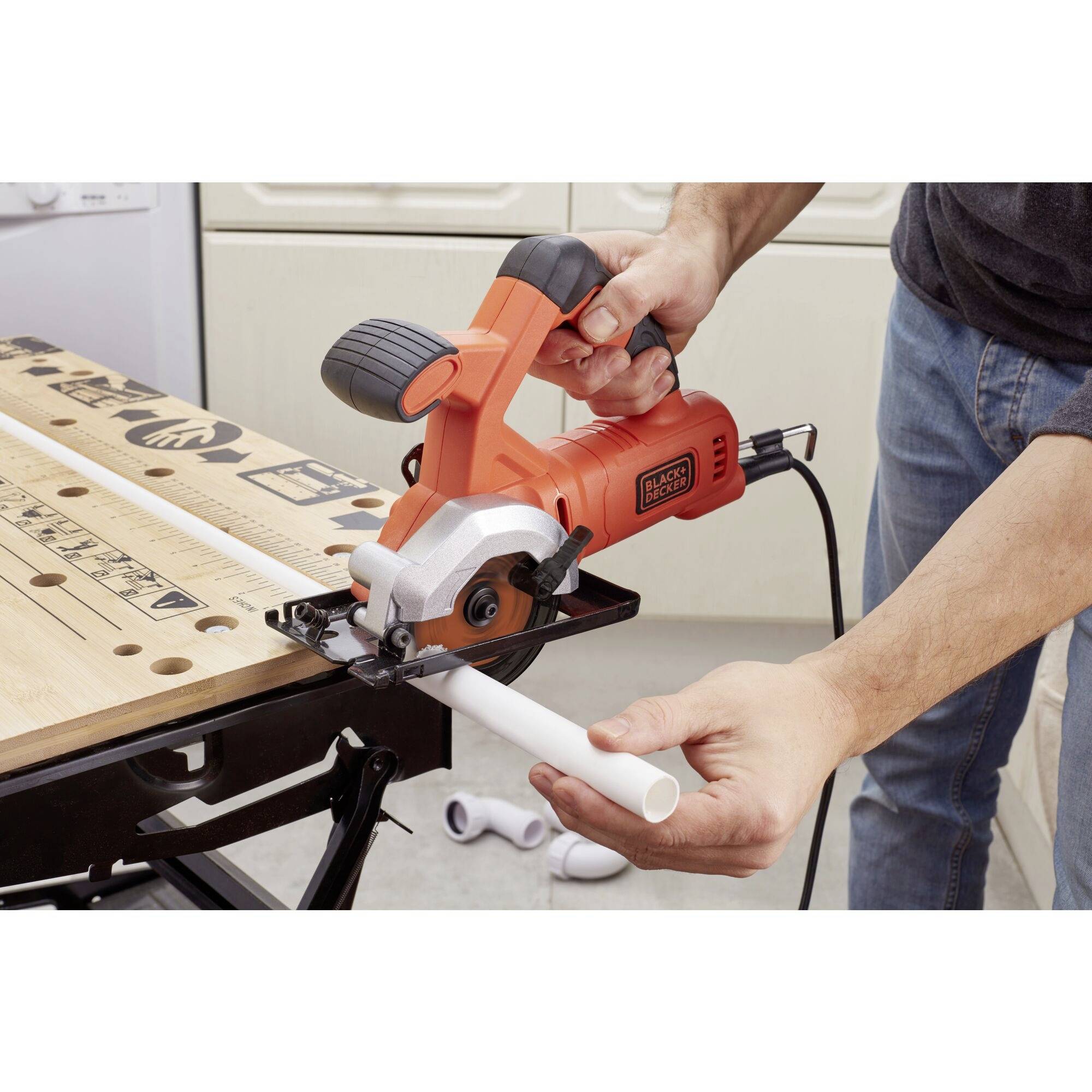 A person is sawing a white pipe with an electric circular hand saw on a workbench. Furniture is visible in the background.