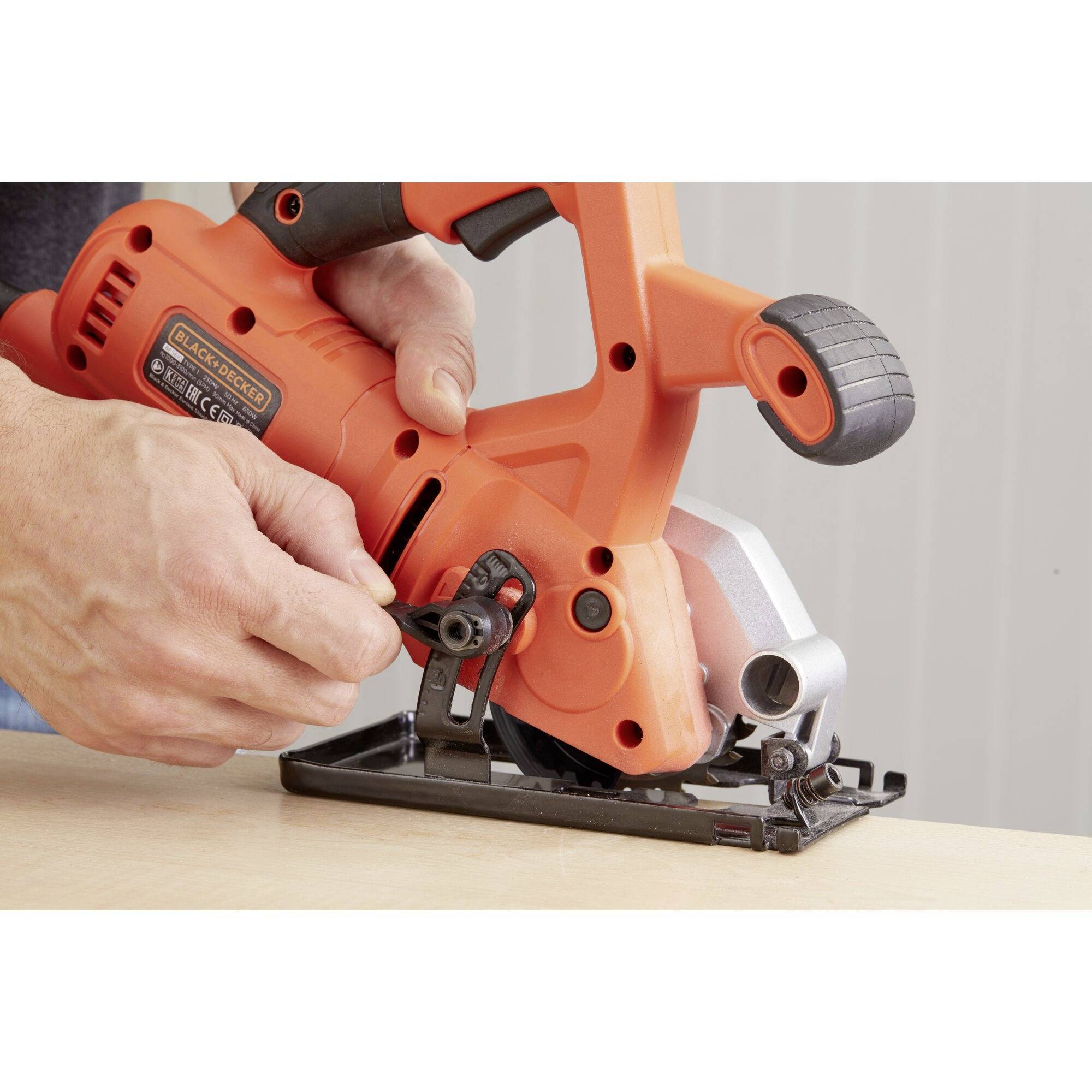 A person adjusts the angle of an orange circular saw on a wooden table to alter the cutting depth.