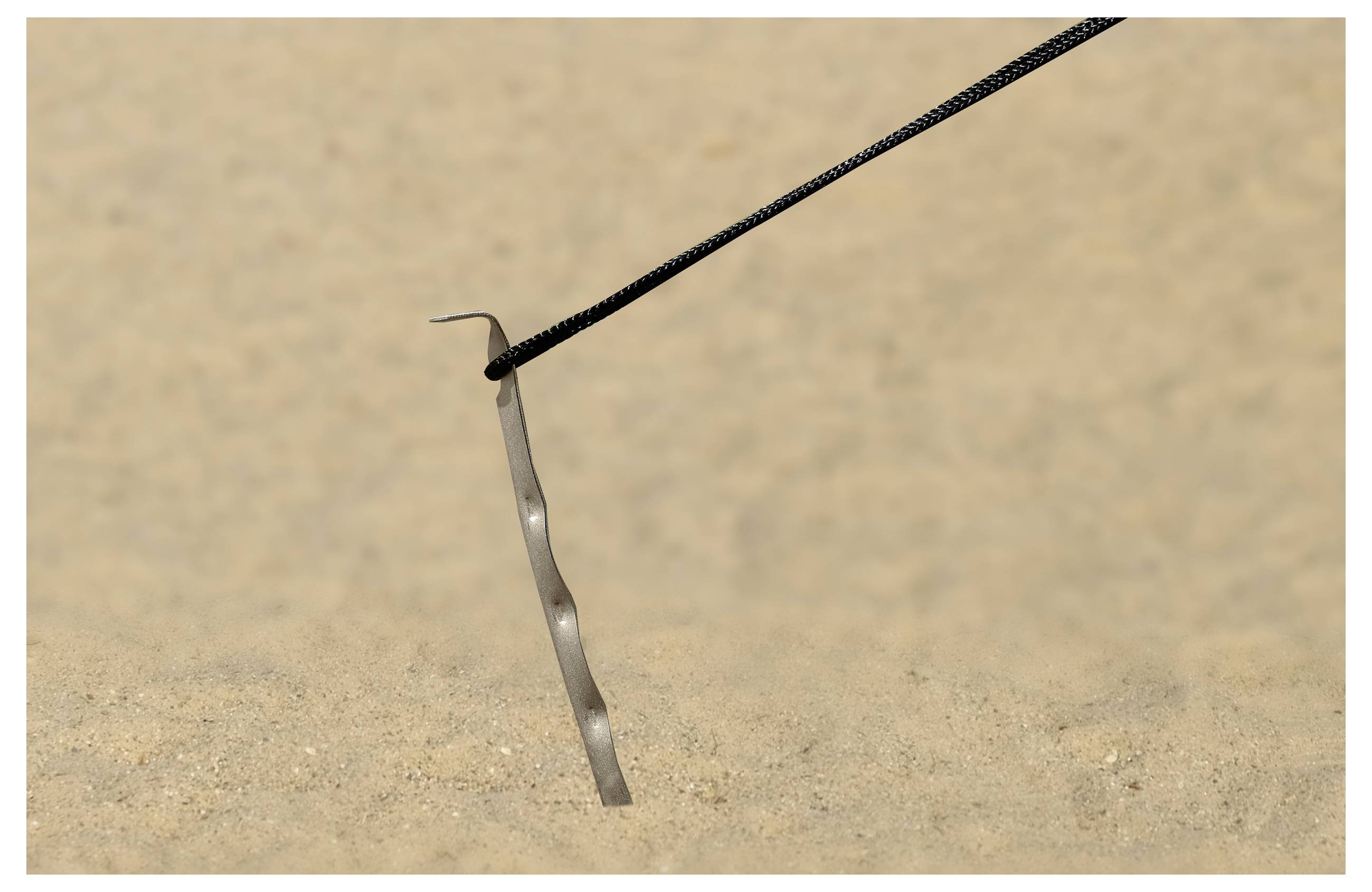 A herring anchor driven into the ground of a sandy terrain. A rope is fastened to the end of the anchor.
