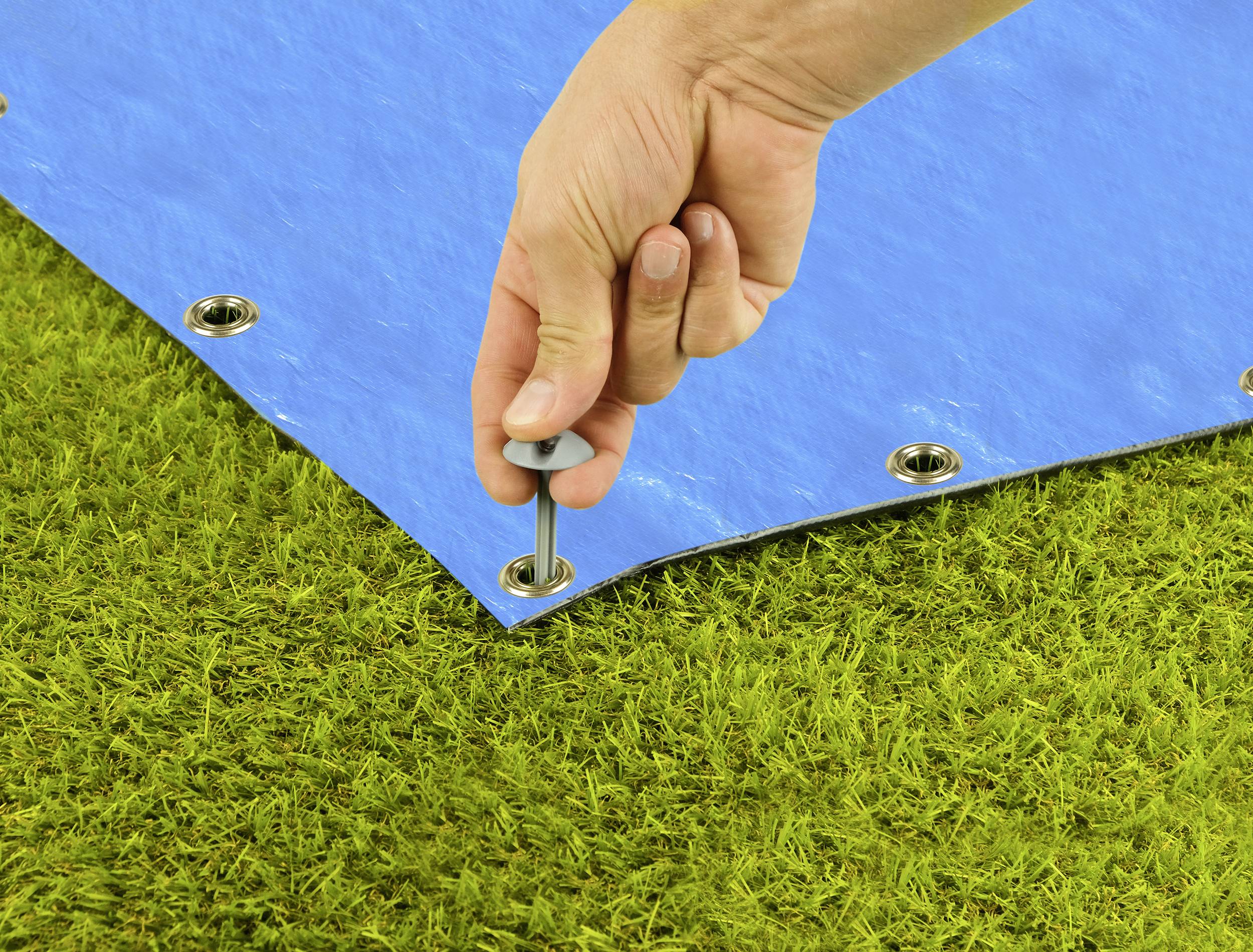 A hand is securing a blue tarpaulin with a bolt on green grass. The tarpaulin has eyelets for fastening.