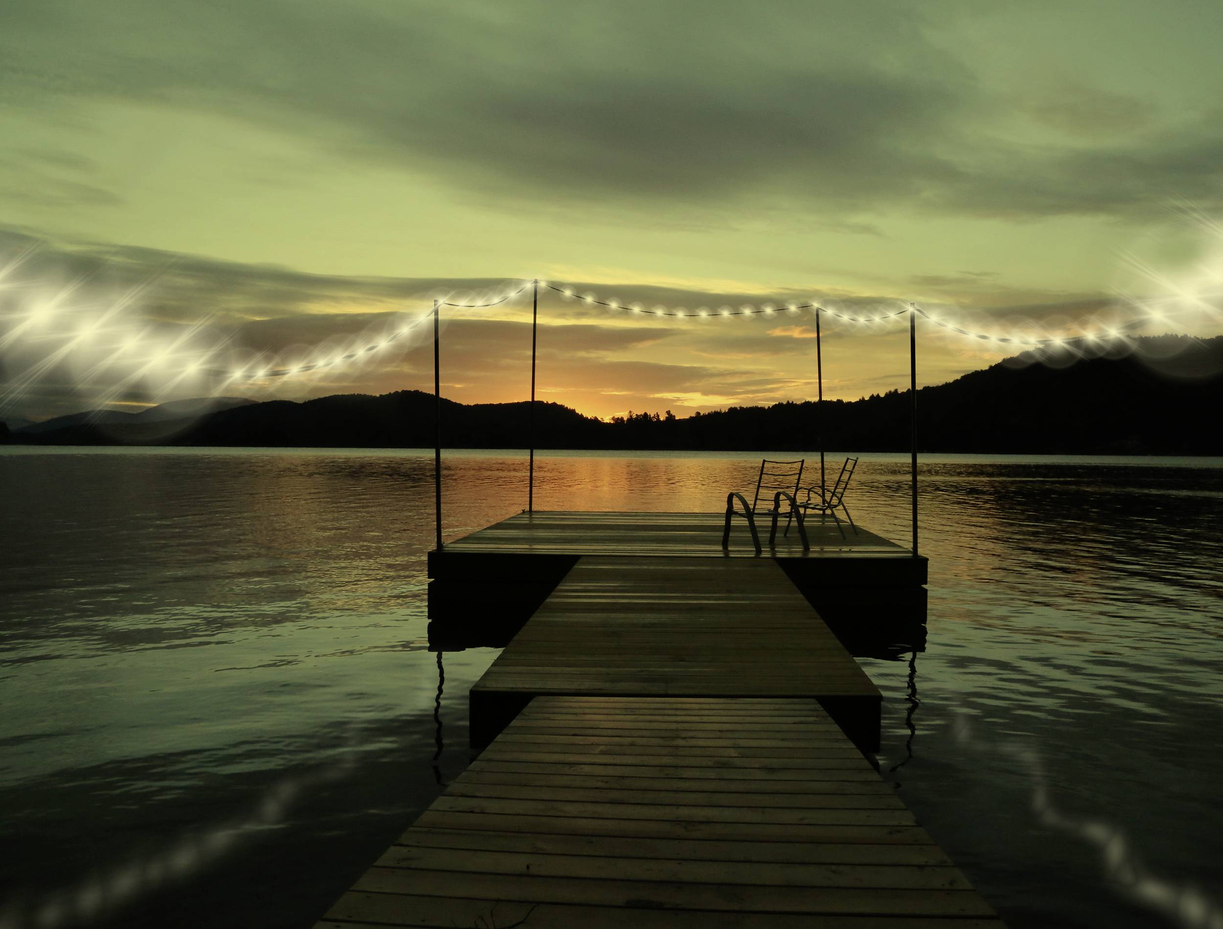 A jetty leads out onto a lake at sunset. Fairy lights hang along its edges. Mountains and a dark sky form the backdrop.