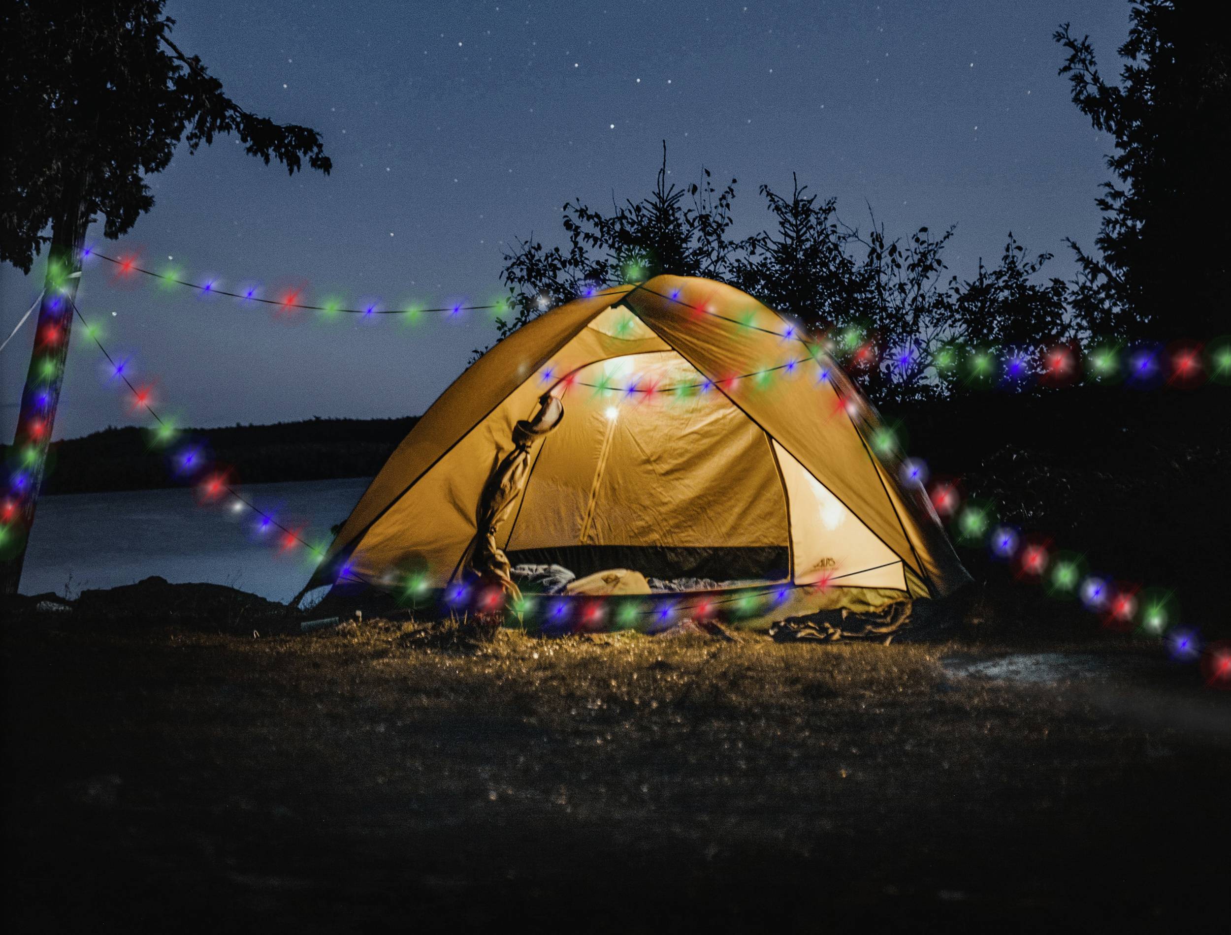 A yellow tent stands illuminated at night by a lakeside, surrounded by colourful fairy lights and stars in the sky.
