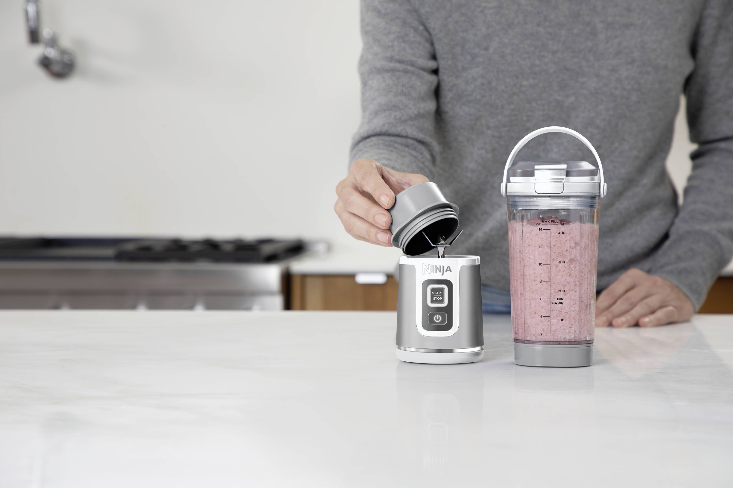 A person is pouring liquid into a small blender. Next to it stands a filled shaker with a red drink. Kitchen in the background.