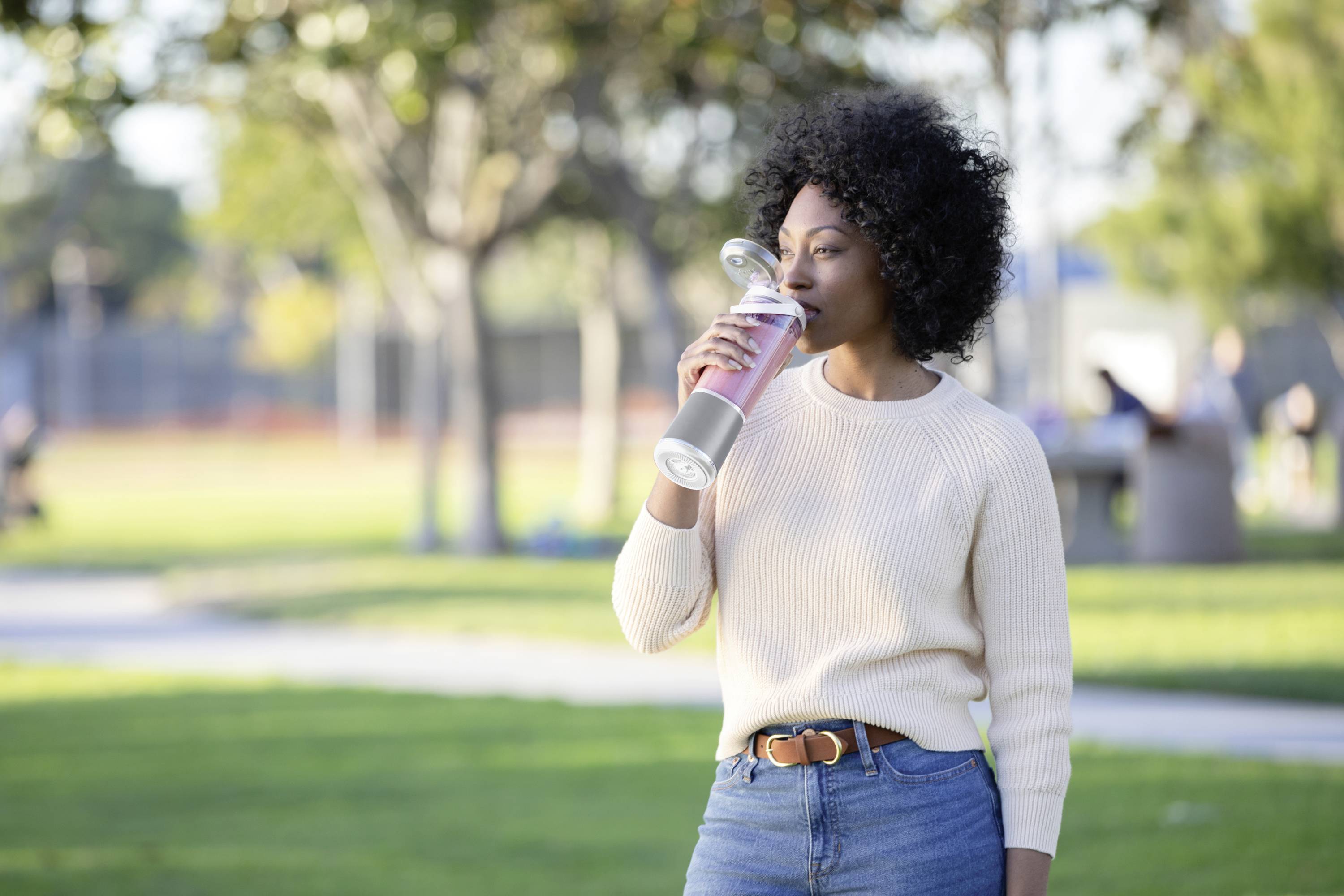 A person is drinking from a large water bottle in a sunny park. Trees and people on grassy areas are visible in the background.