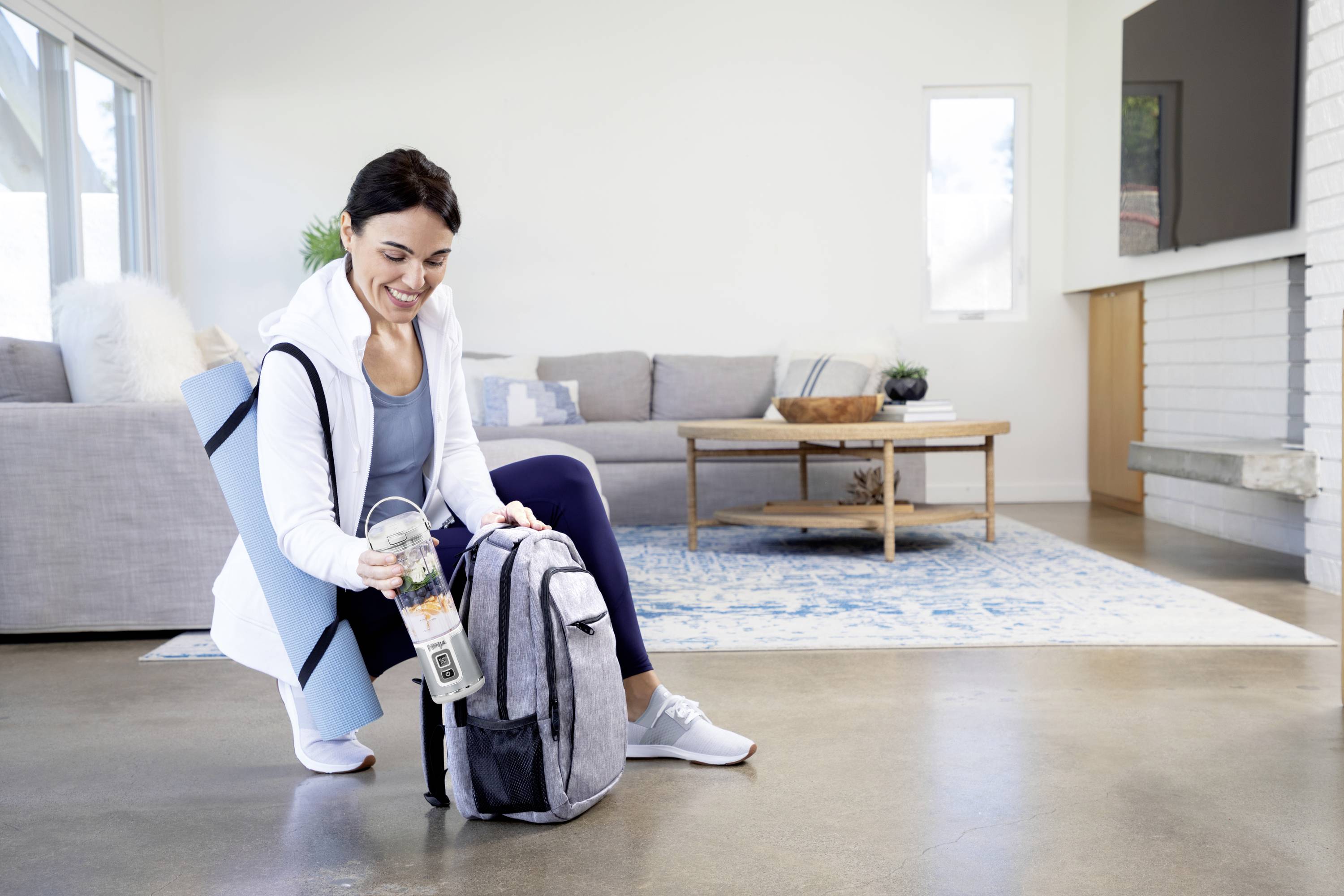 A woman is sitting on the floor of a living room, packing a grey bag and holding a water bottle. A yoga mat is lying beside her.