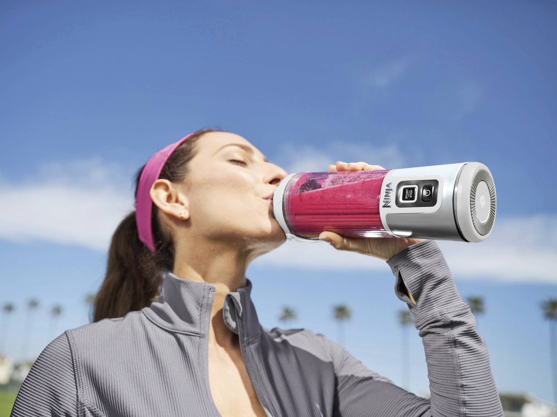 A woman is drinking from a portable blender containing a red smoothie. Blurred palm trees are visible in the background.