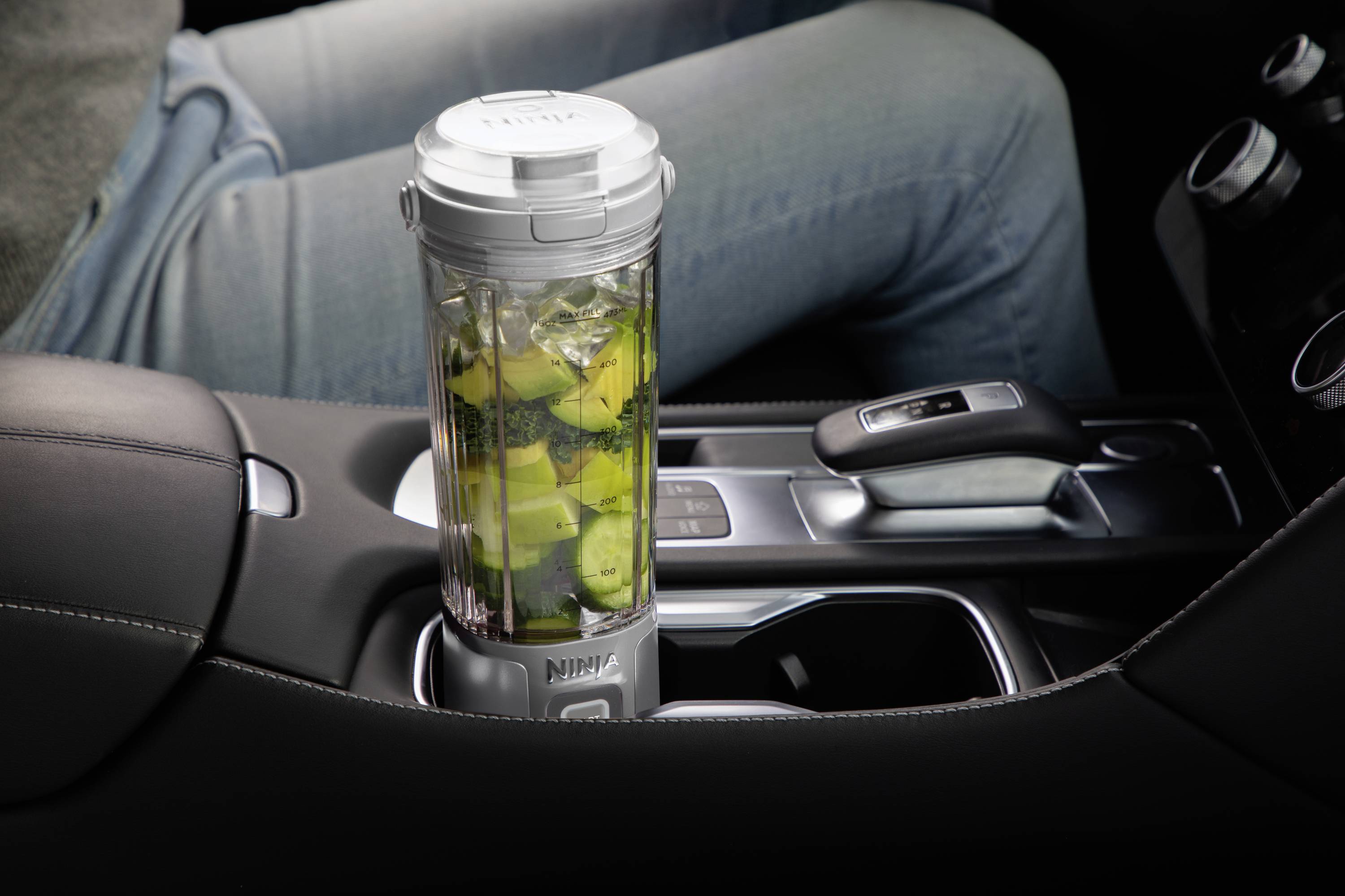 A portable blender filled with fruit and vegetables sits in the centre console of a car. A person is seated at the wheel.