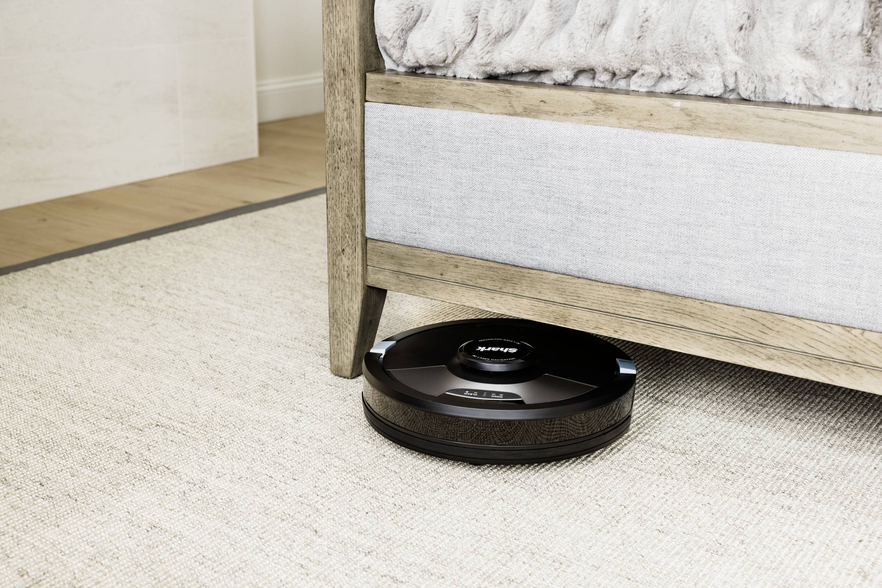 A vacuum robot is cleaning the floor beneath a bed in a brightly furnished room.