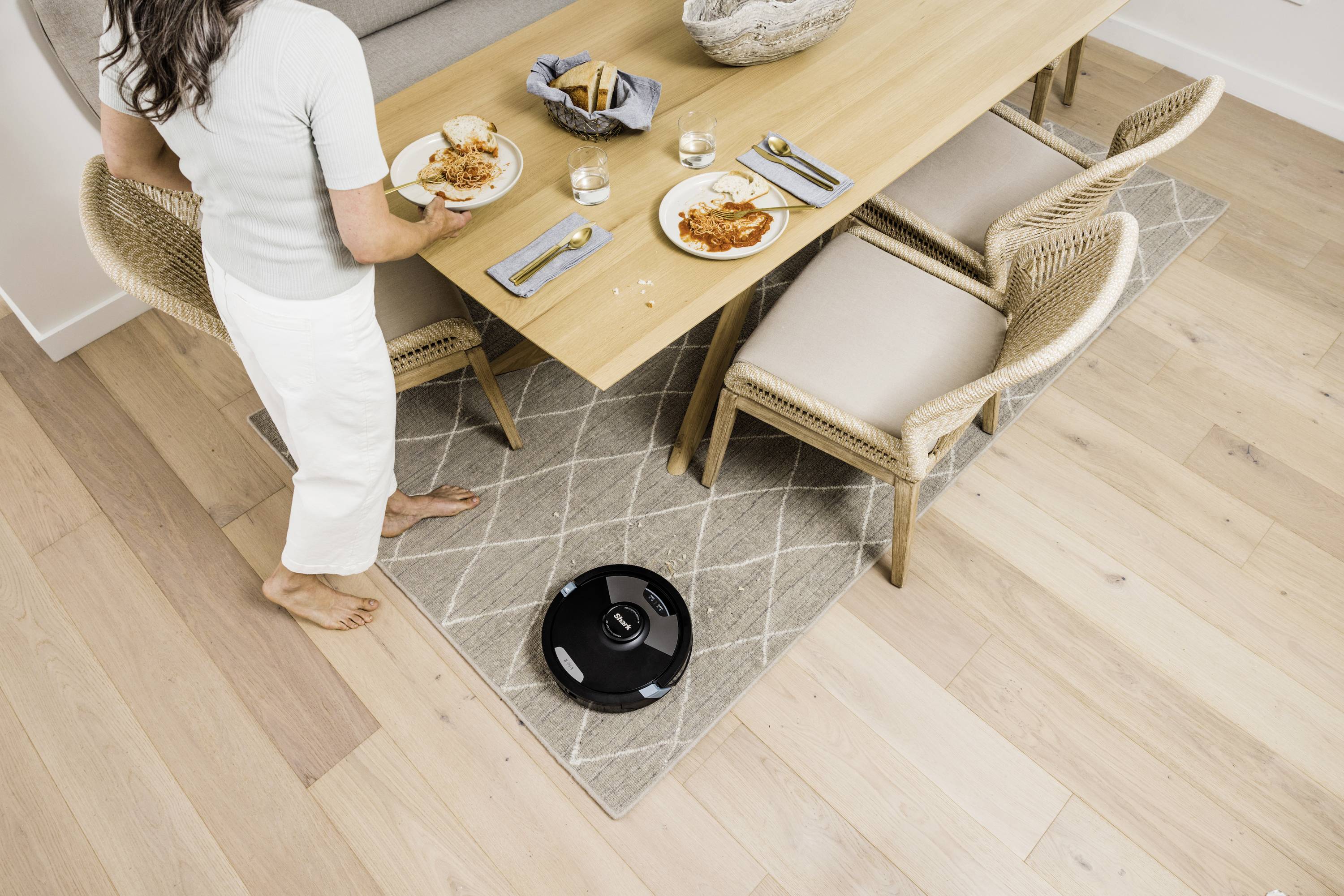 A person is standing with a plate next to a laid dining table, while a black robotic vacuum cleaner is cleaning the floor.