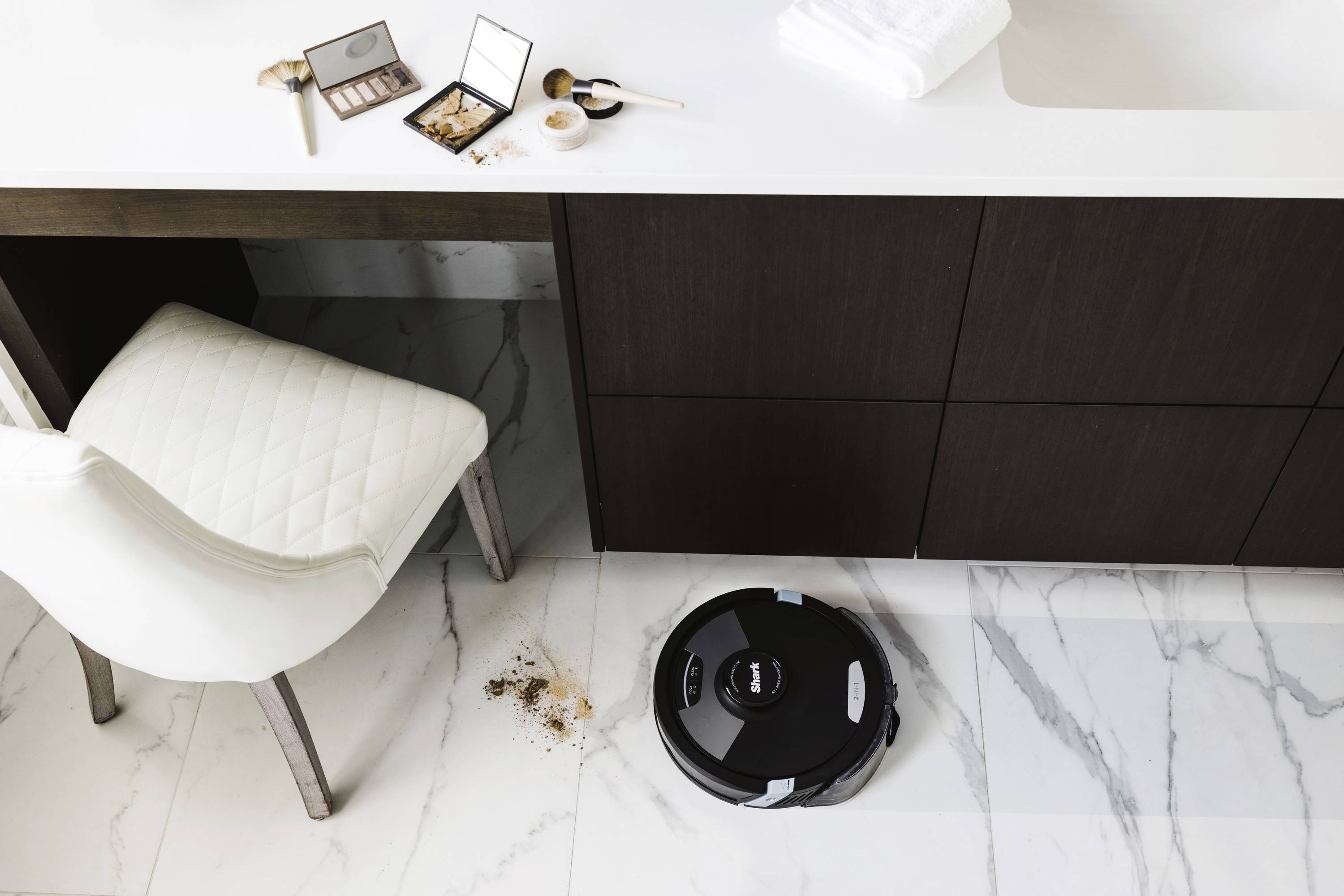 A robotic vacuum cleaner is cleaning the floor beneath a modern washbasin. Cosmetics are scattered on the worktop. Clean, minimalist bathroom.