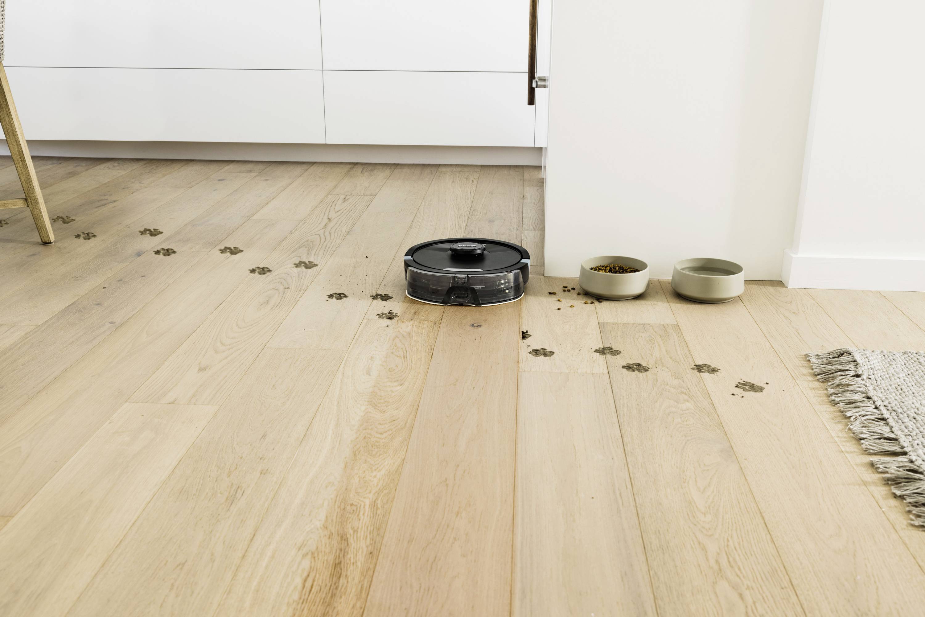 A robot vacuum is cleaning a wooden floor with muddy tracks in a kitchen. Two pet food bowls are placed beside it.