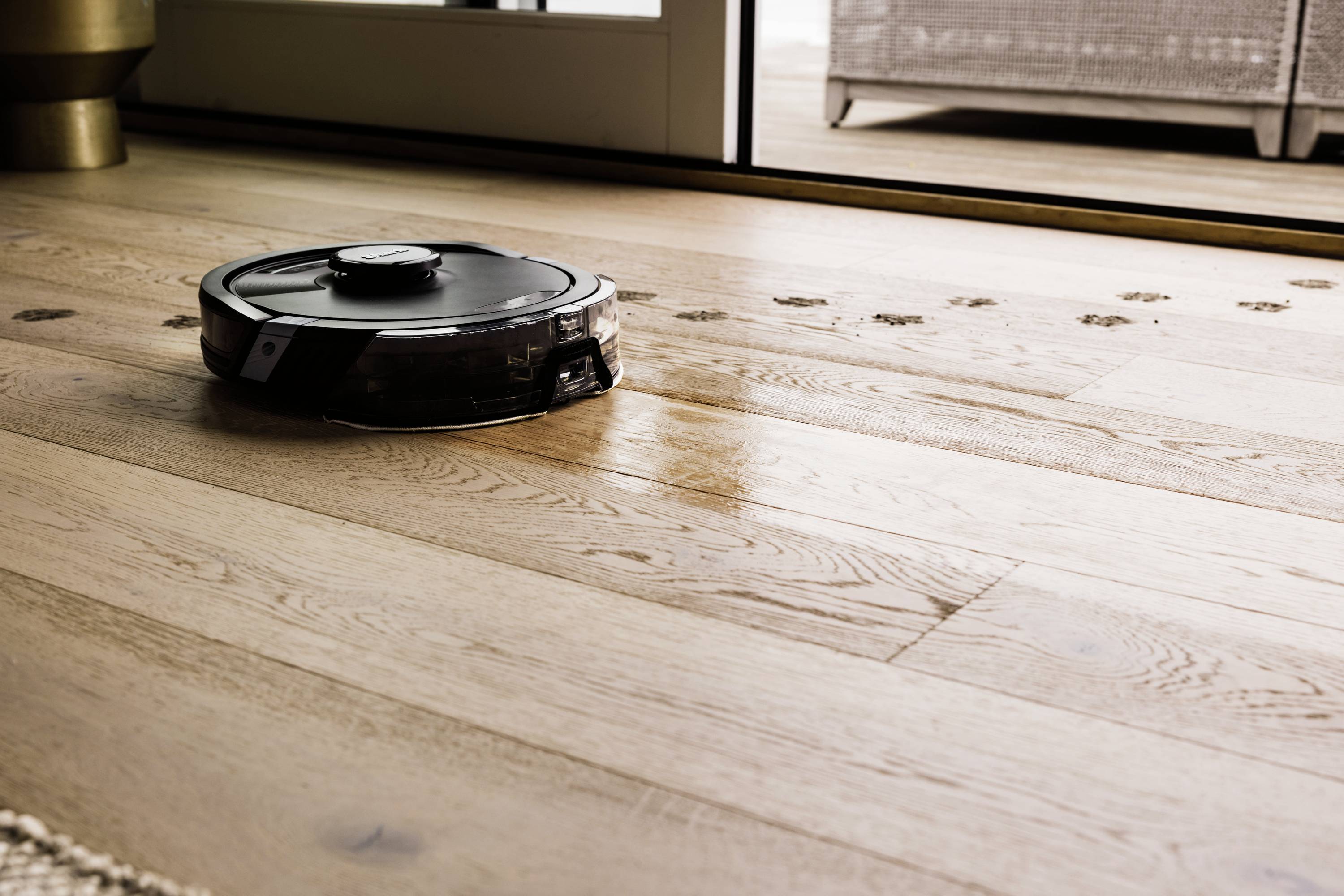 A robot vacuum cleaner is cleaning wooden floors in a bright room. It removes dirt tracks leading towards a glass door.