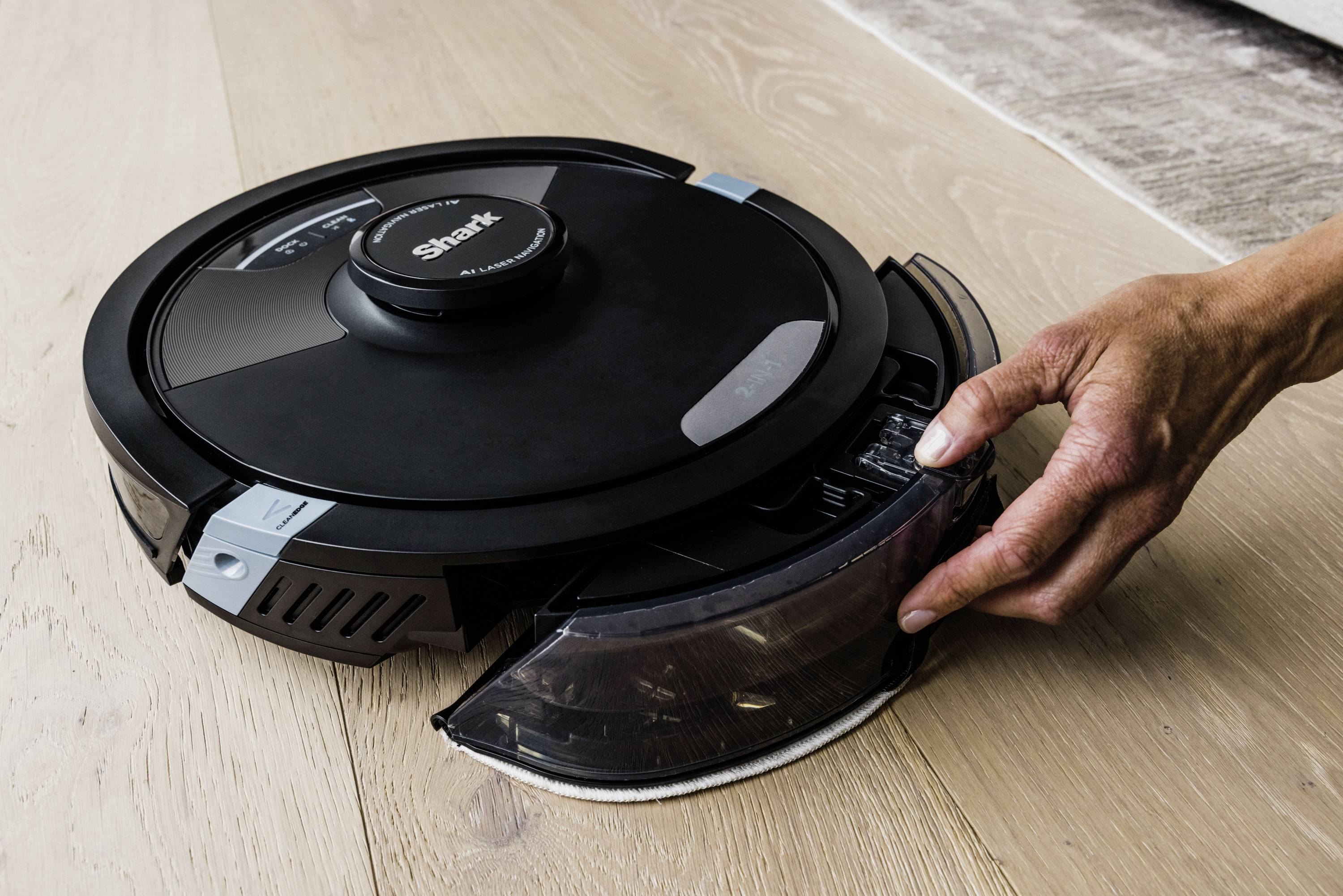 A hand removes the dust container of a round robot vacuum on a wooden floor.