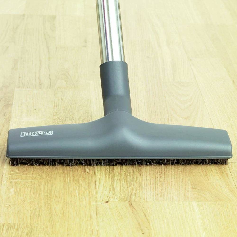 Vacuum cleaner nozzle on a wooden floor. The nozzle has black bristles and a silver handle. The floor is clean and glossy.