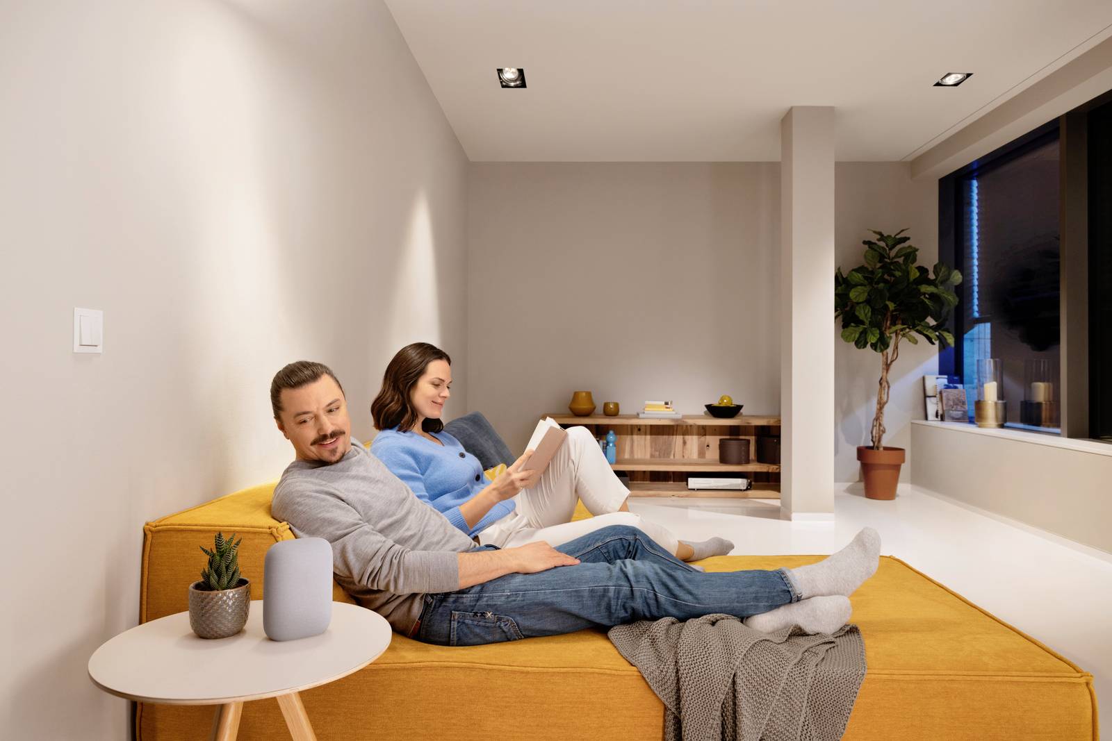 A man and a woman are relaxing on a yellow sofa in a modern living room. Both are holding a tablet and a book.