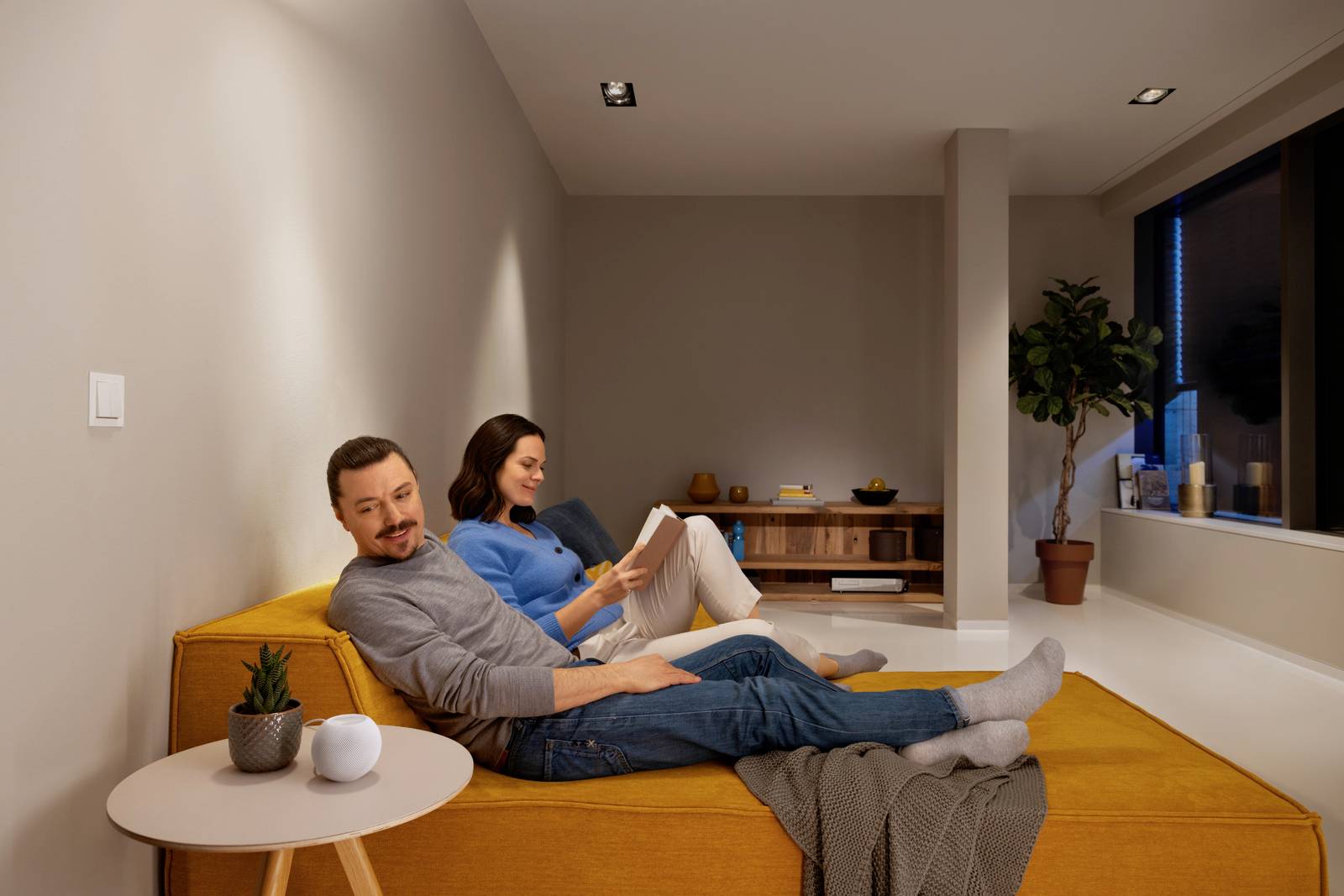 A man and a woman are sitting relaxed on a light-coloured sofa in a modern living room. Both are reading or relaxing.