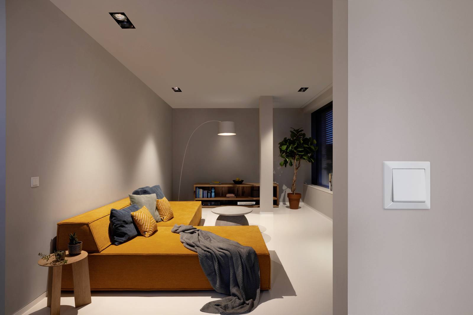 A modern living room with an orange sofa, blue cushions, a standard lamp, and a plant. A light switch is visible on the wall.