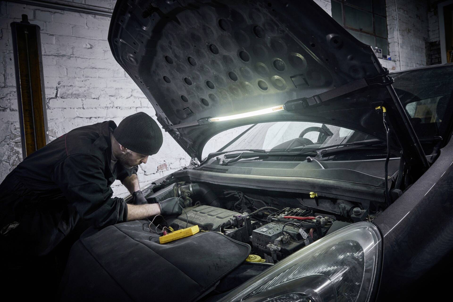A mechanic in a workshop, working underneath the raised bonnet of a car. The room is dimly lit.