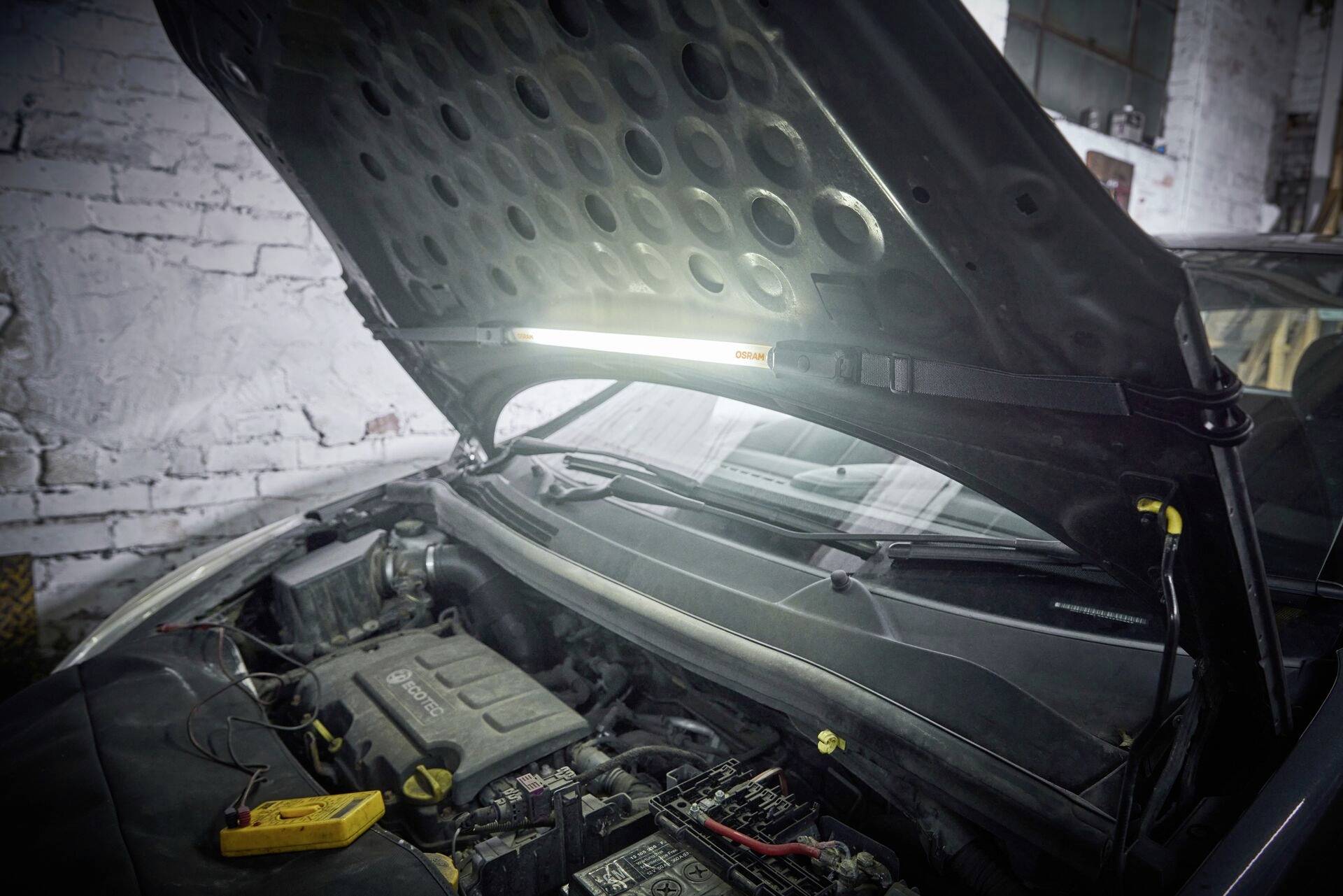 A car is parked with its bonnet open in a garage workshop. A diagnostic device is lying next to the engine to support maintenance work.