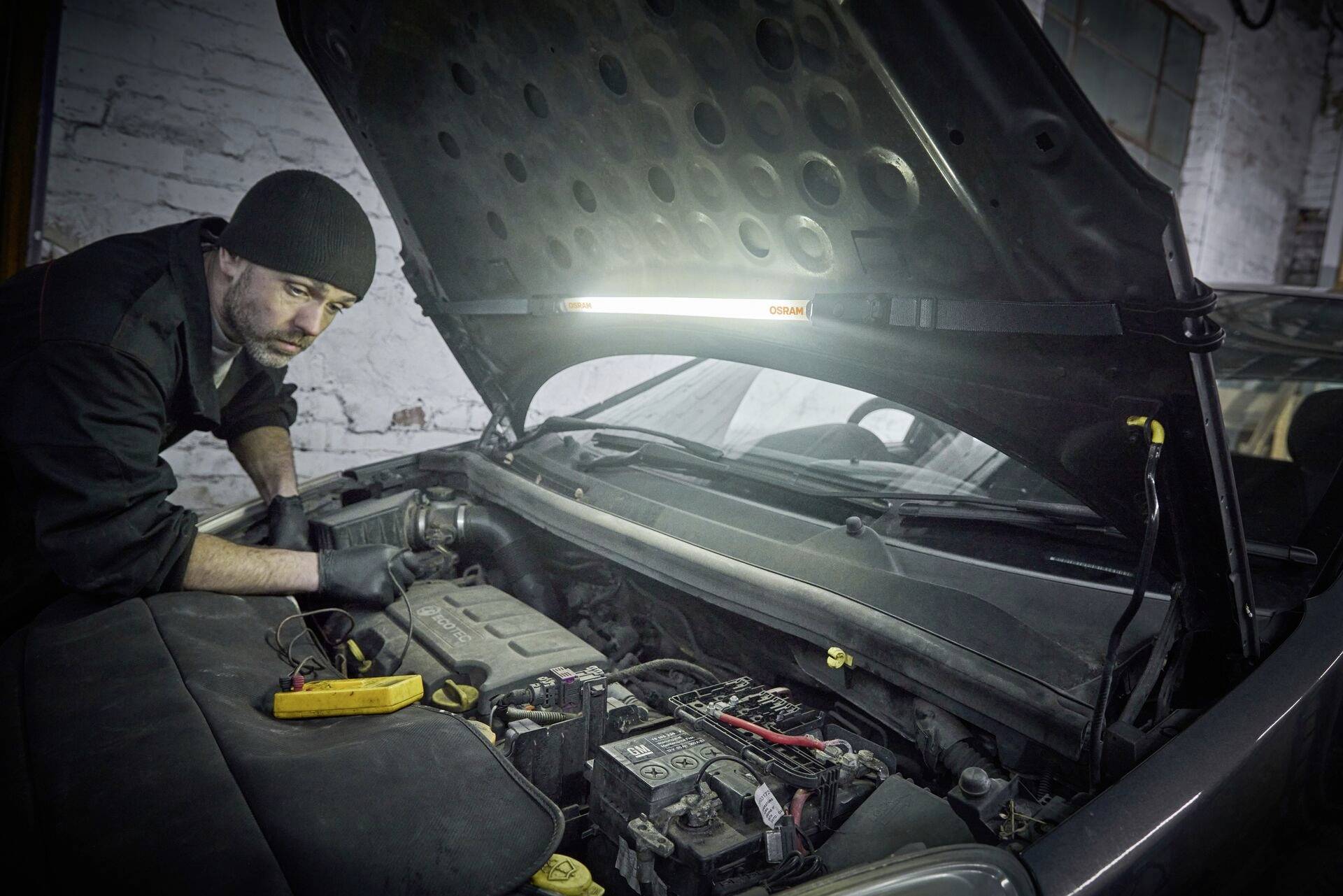 A mechanic in dark clothing is working with a measuring instrument in an open engine compartment of a car within a workshop environment.
