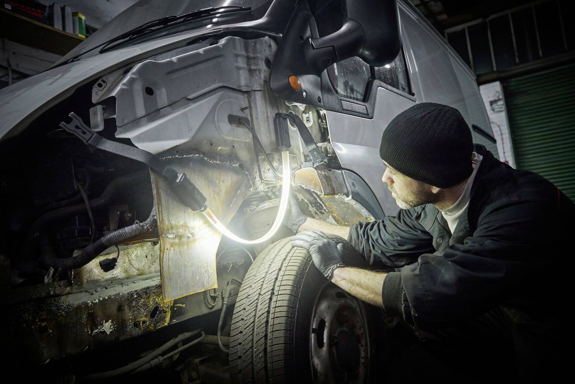 A mechanic inspects the damaged wing of a van in a workshop. The lighting reveals clear rust marks on the vehicle's chassis.