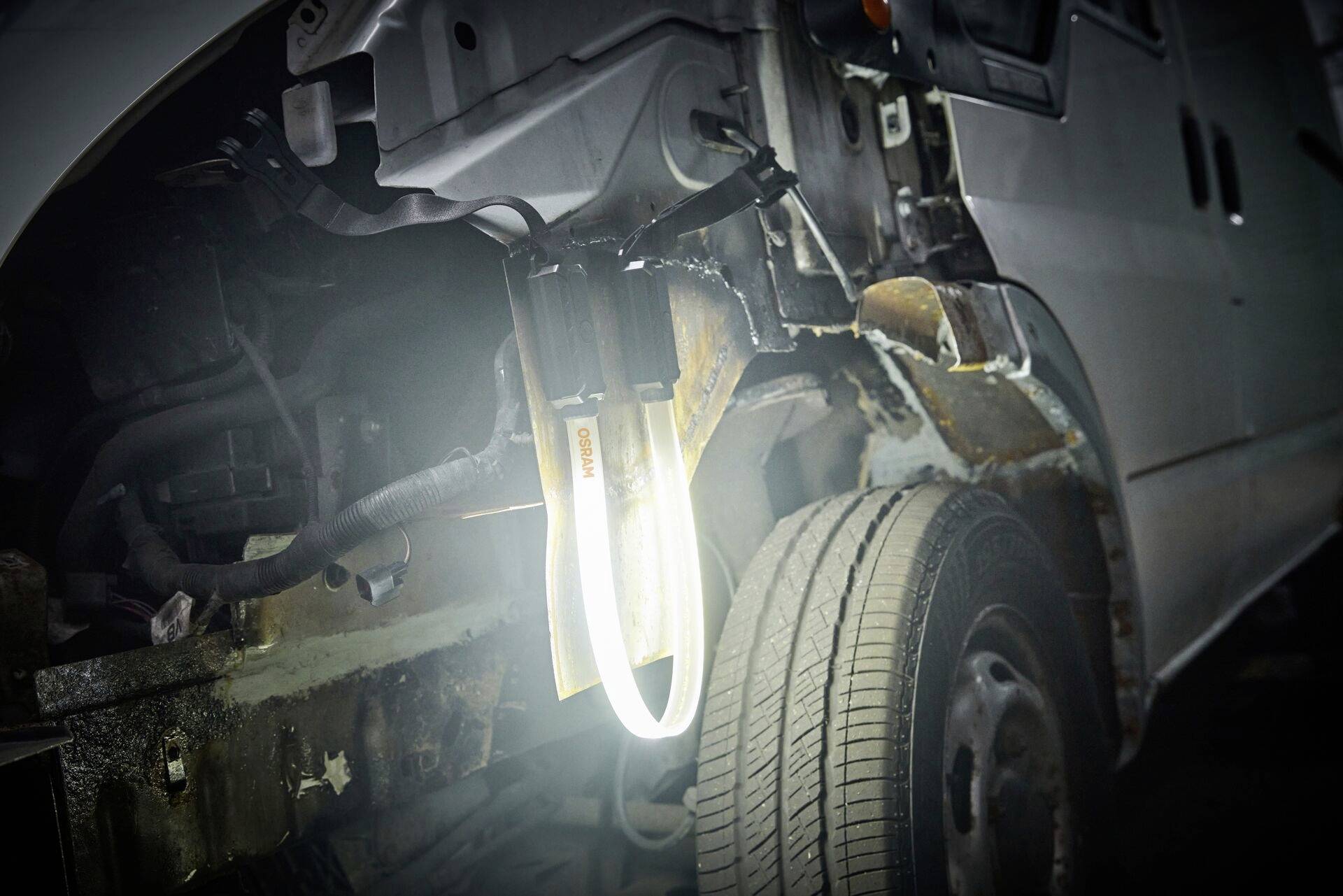 Ein Auto mit geöffneter Motorhaube zeigt einen rostigen Fahrzeugrahmen und sichtbare Verkabelung, beleuchtet von einer Werkstattlampe.