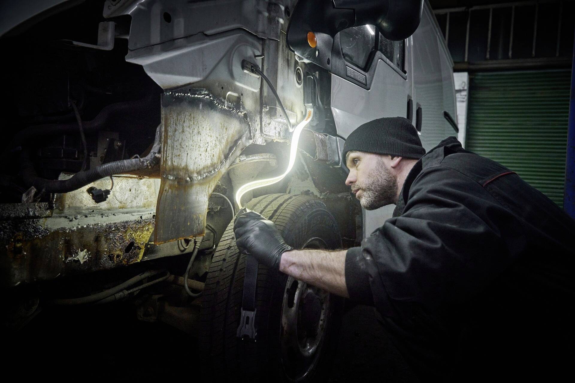 A mechanic in workwear is inspecting the interior of a car with an inspection lamp, focused on a rusted component.