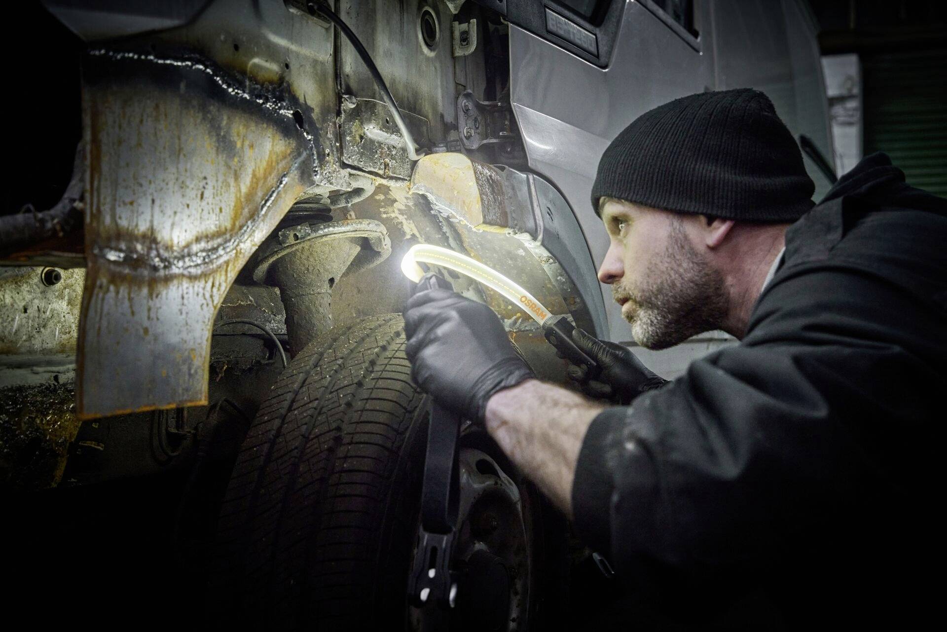 A mechanic carefully inspects the rusty wheel arch of a vehicle in a workshop. He is using a torch for better visibility.
