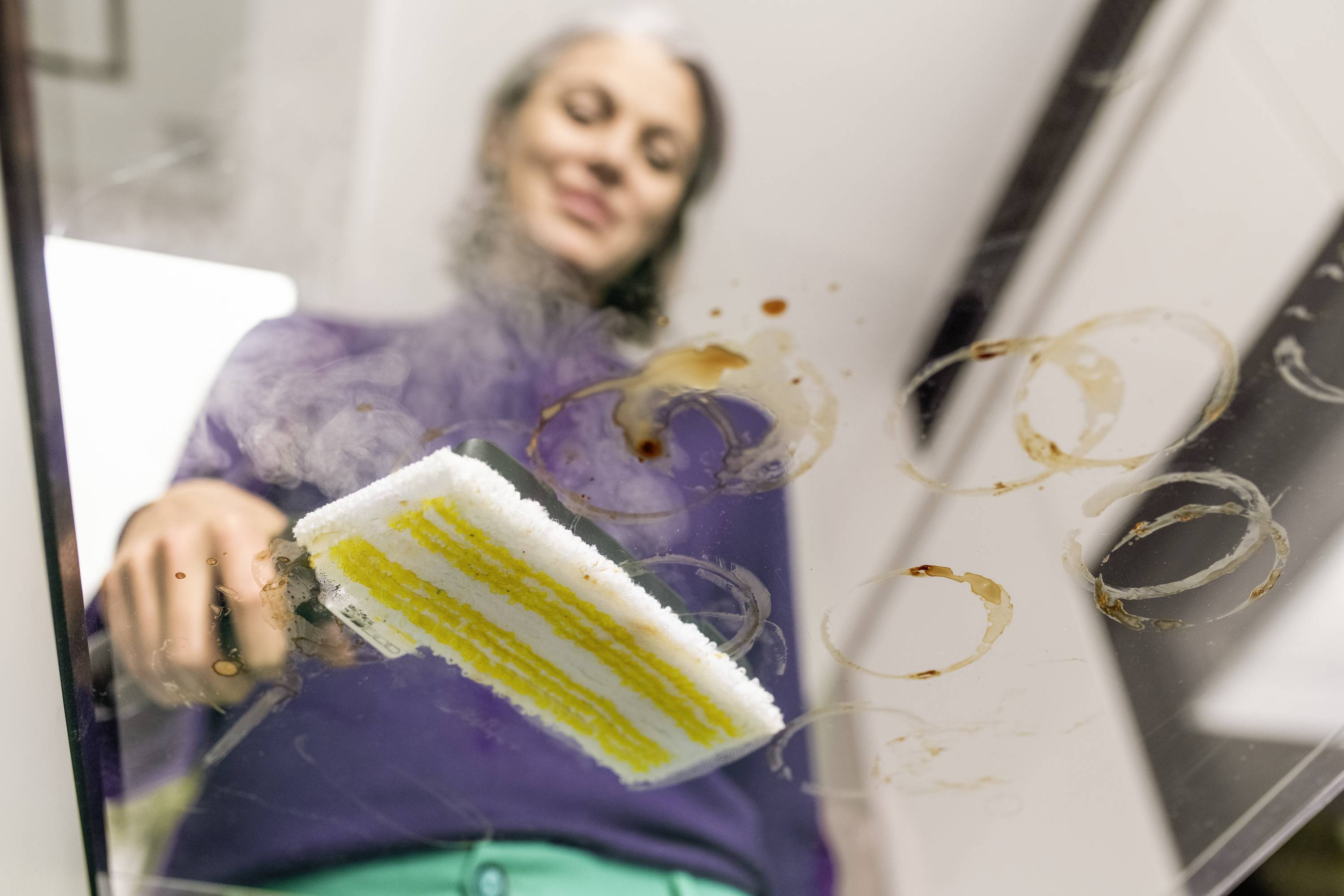 A person is cleaning a dirty glass surface with a sponge, viewed from a perspective underneath the glass.