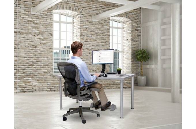 A person is sitting at a desk in a modern office with brick walls, working on a computer, with plants and windows in the background.