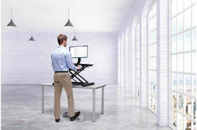A man is standing at an adjustable-height desk in a modern, bright office with large windows, using a computer.