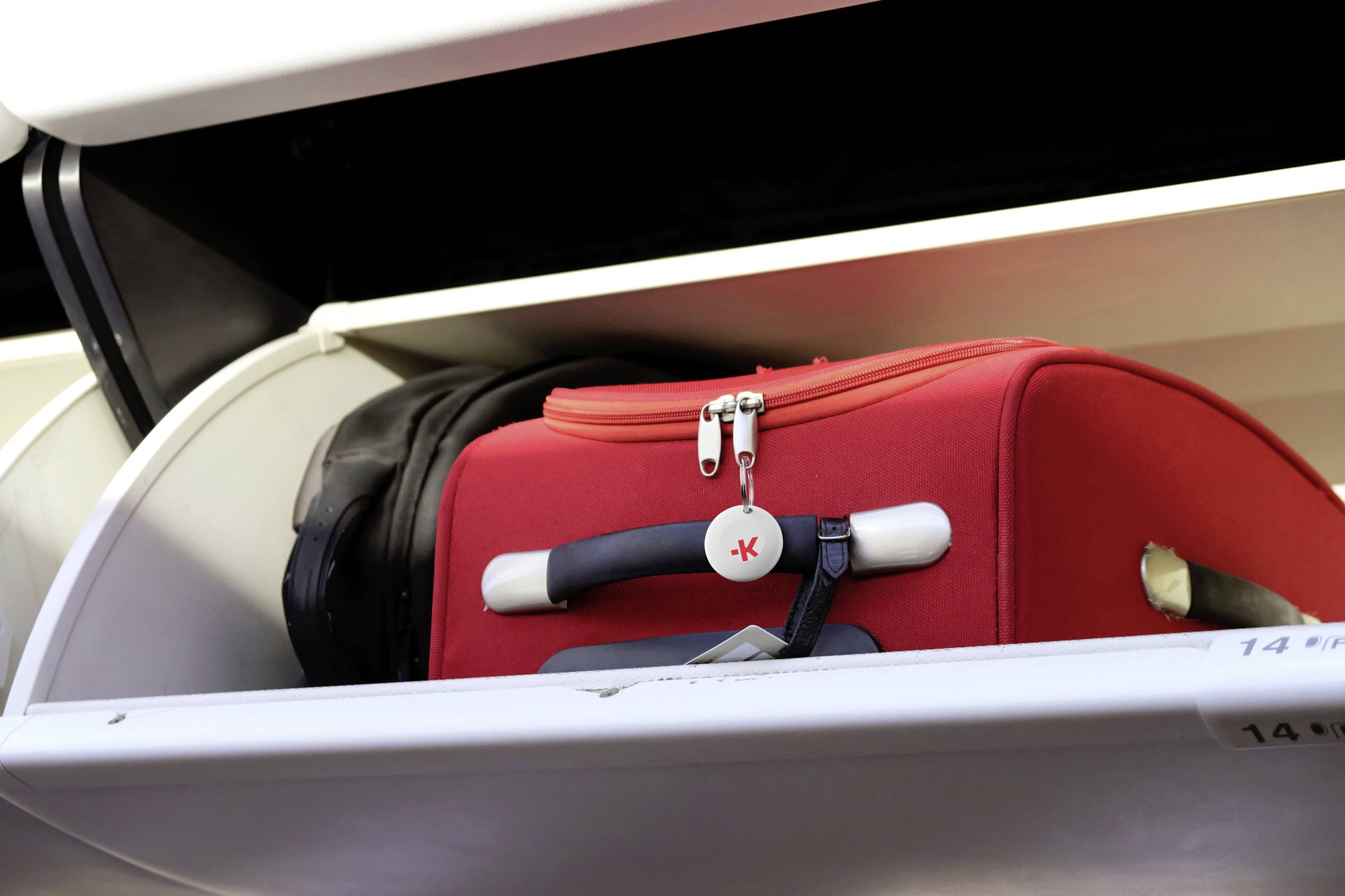 Red suitcase with label stowed in an aircraft overhead locker. A folded jacket lies beside it.