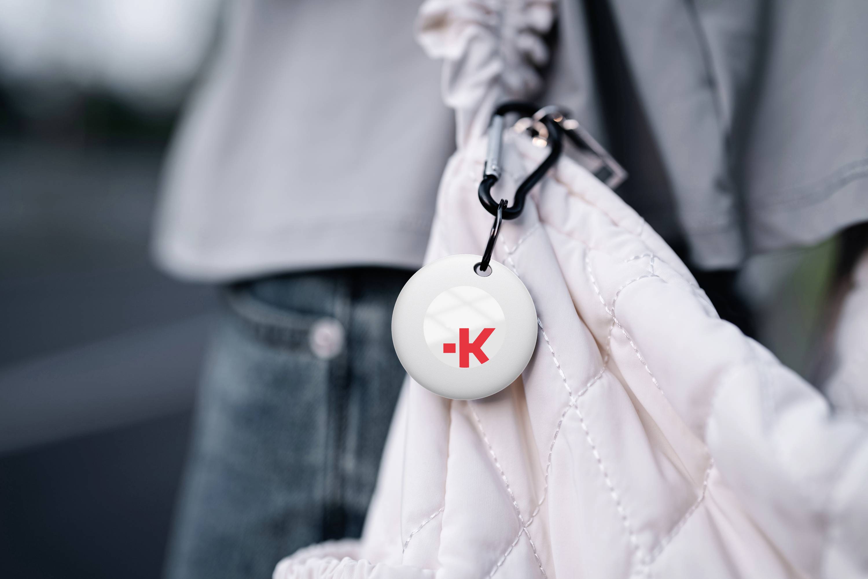 A keyring with a red 'K' logo hangs from a white, quilted bag. The person is wearing a grey jacket.