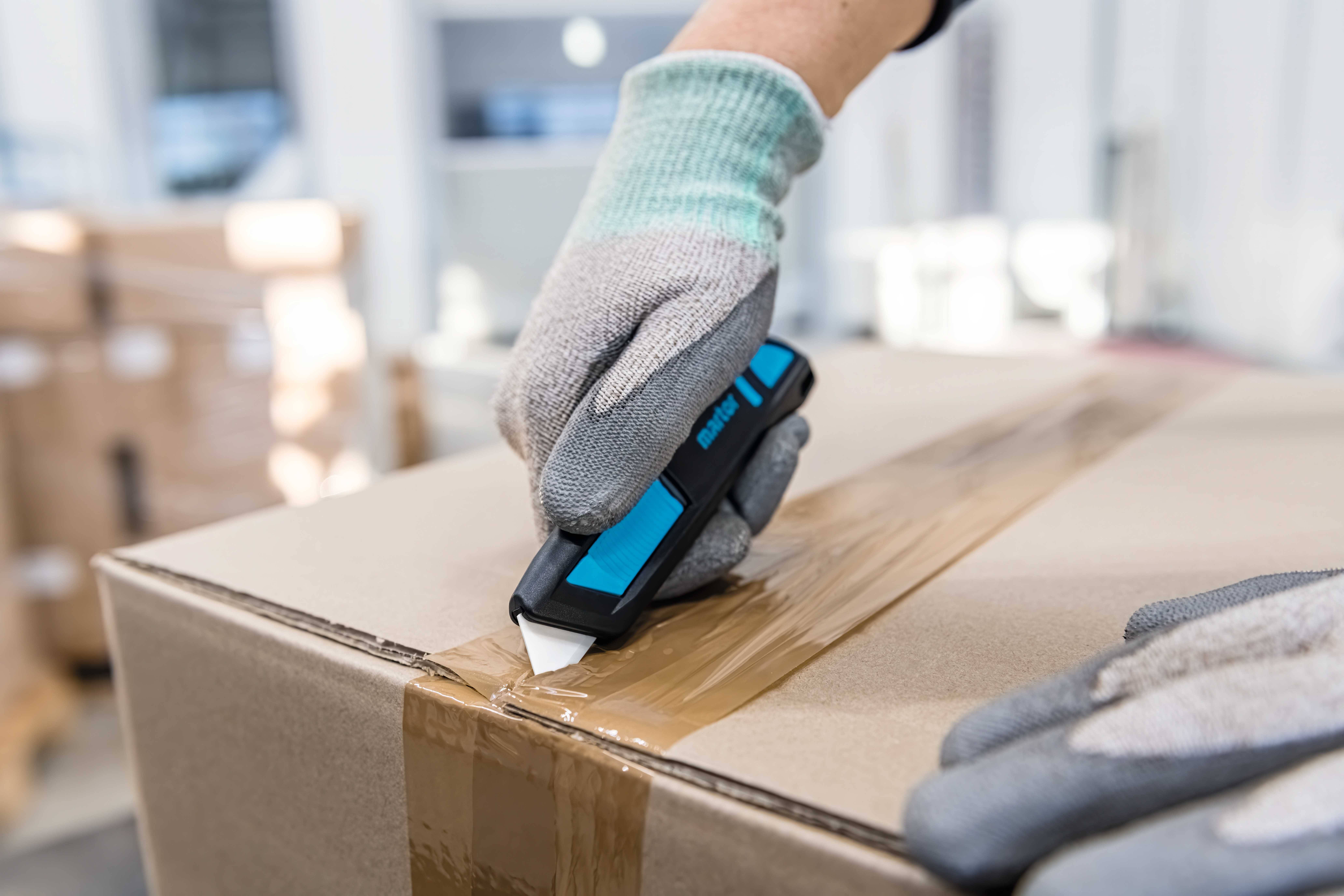 A person wearing work gloves is cutting tape on a cardboard box with a blue utility knife in a warehouse.