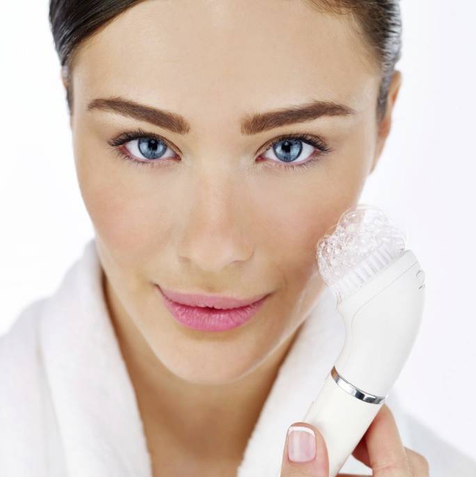 A woman in a dressing gown is cleansing her face with an electric facial brush. Her facial expression is friendly and relaxed.