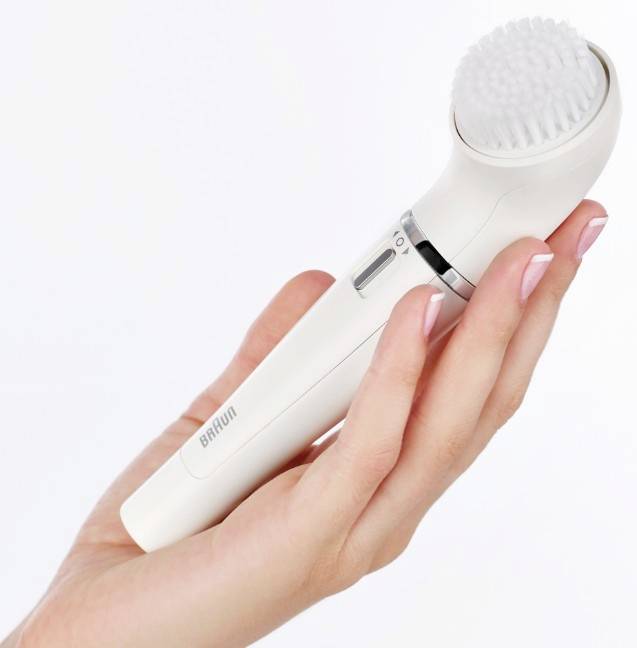 A hand is holding a white electric facial cleansing brush with soft bristles.